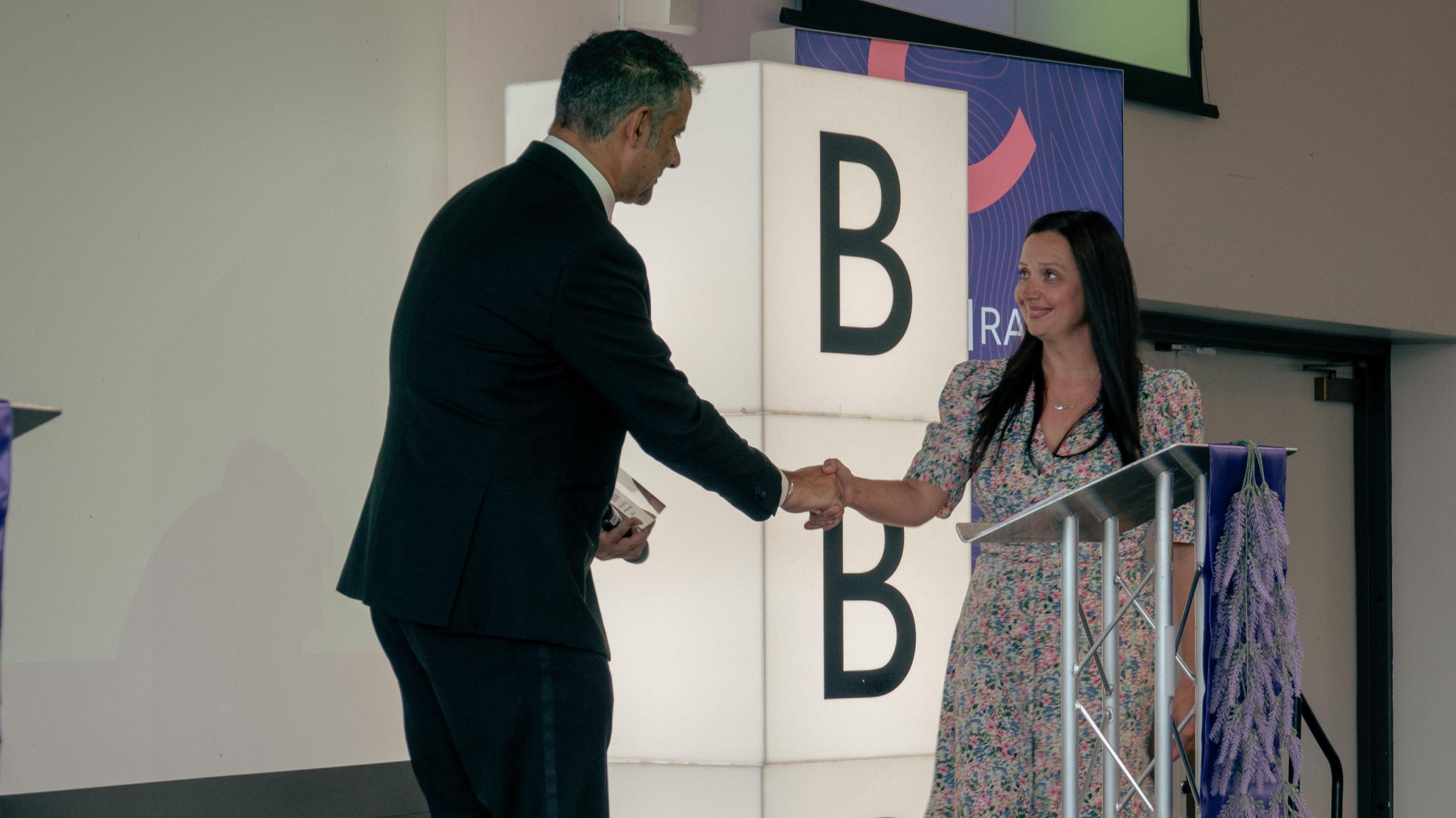 A man shakes a woman's hand in front of a purple plinth