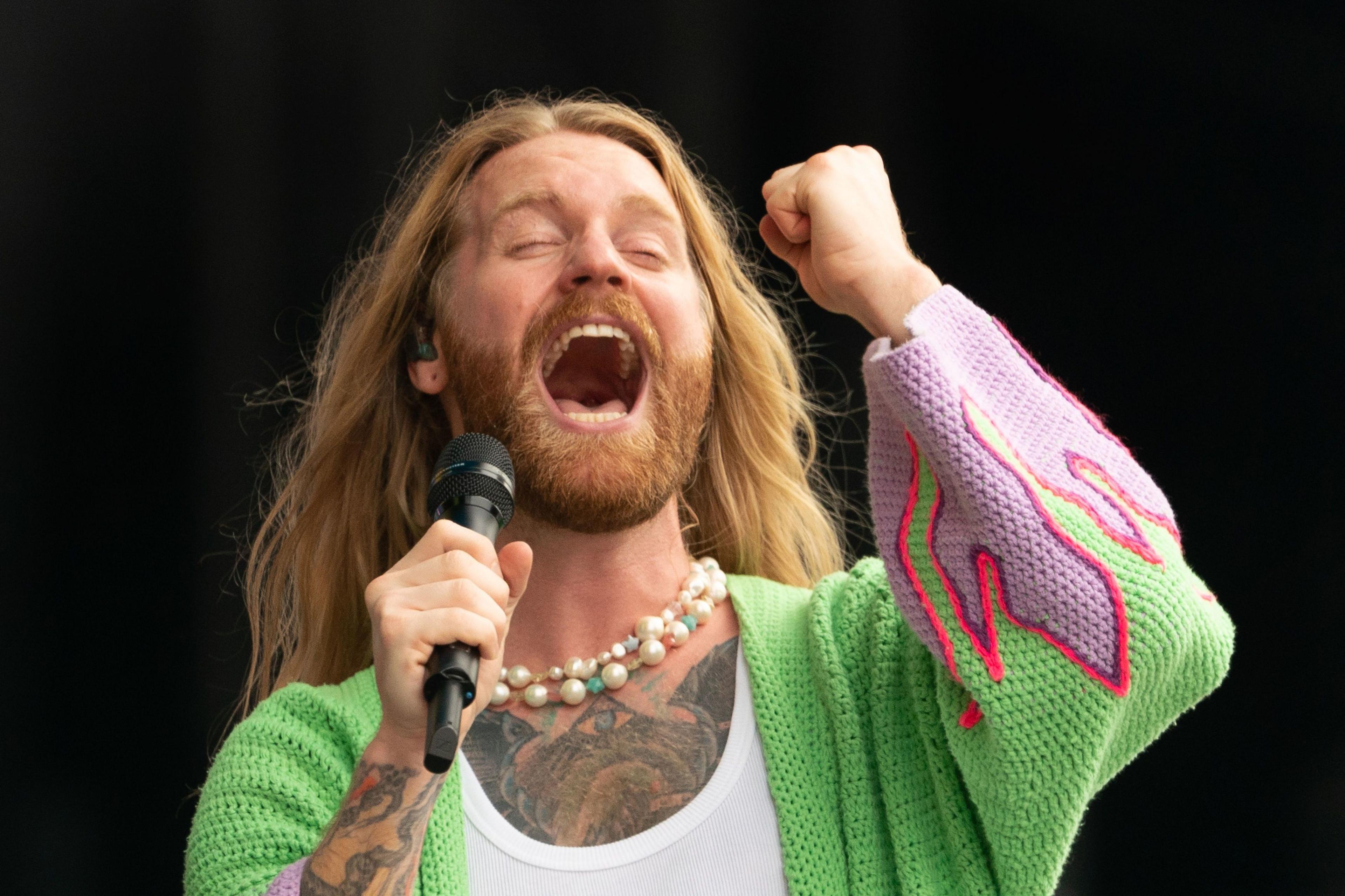 Sam Ryder on stage. He's wearing a green knitted cardigan with purple flame effects on the sleeves; and closes his eyes as he sings an intense note.