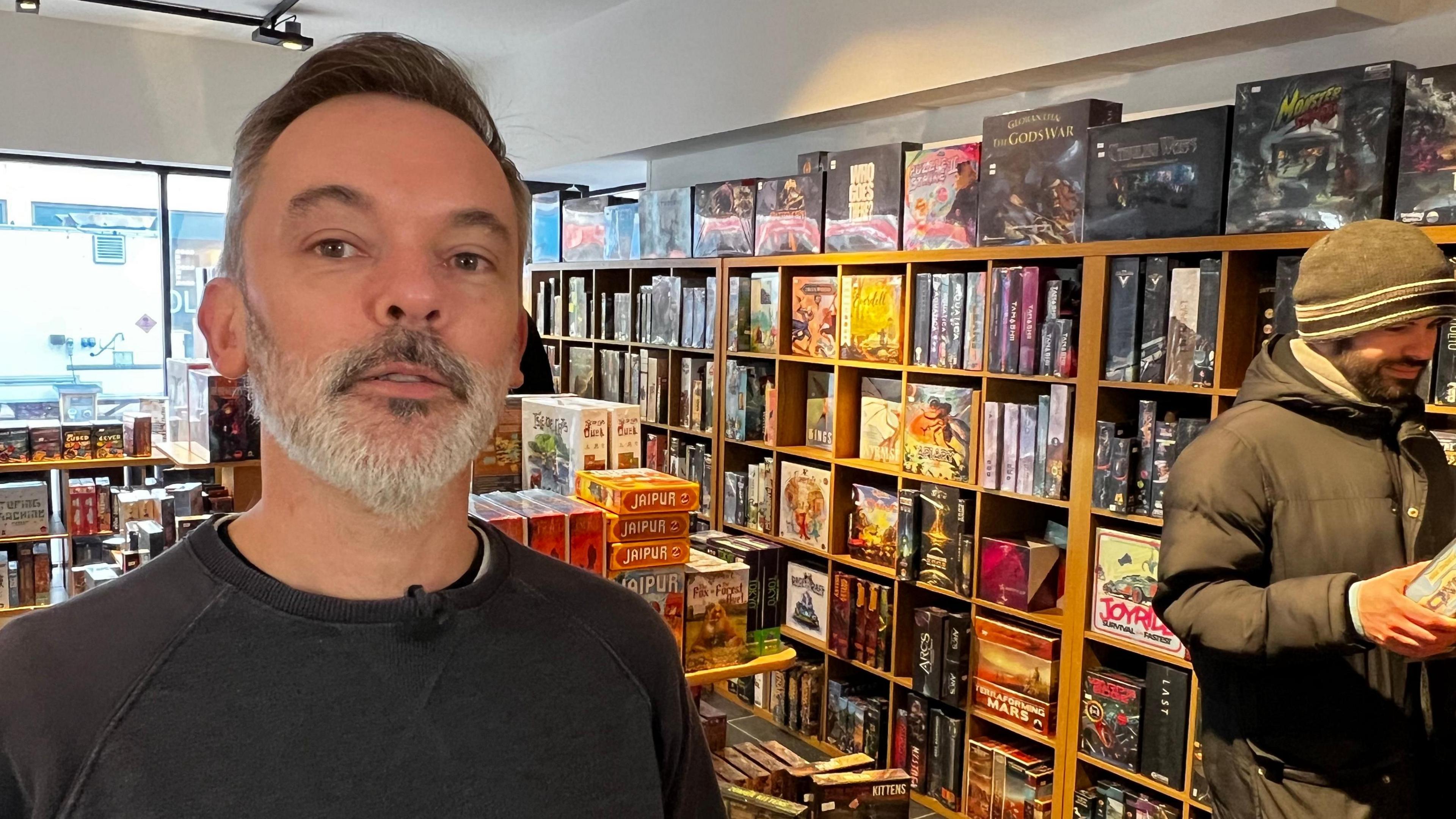 Man wearing black jumper stood in front of board games inside a shop.