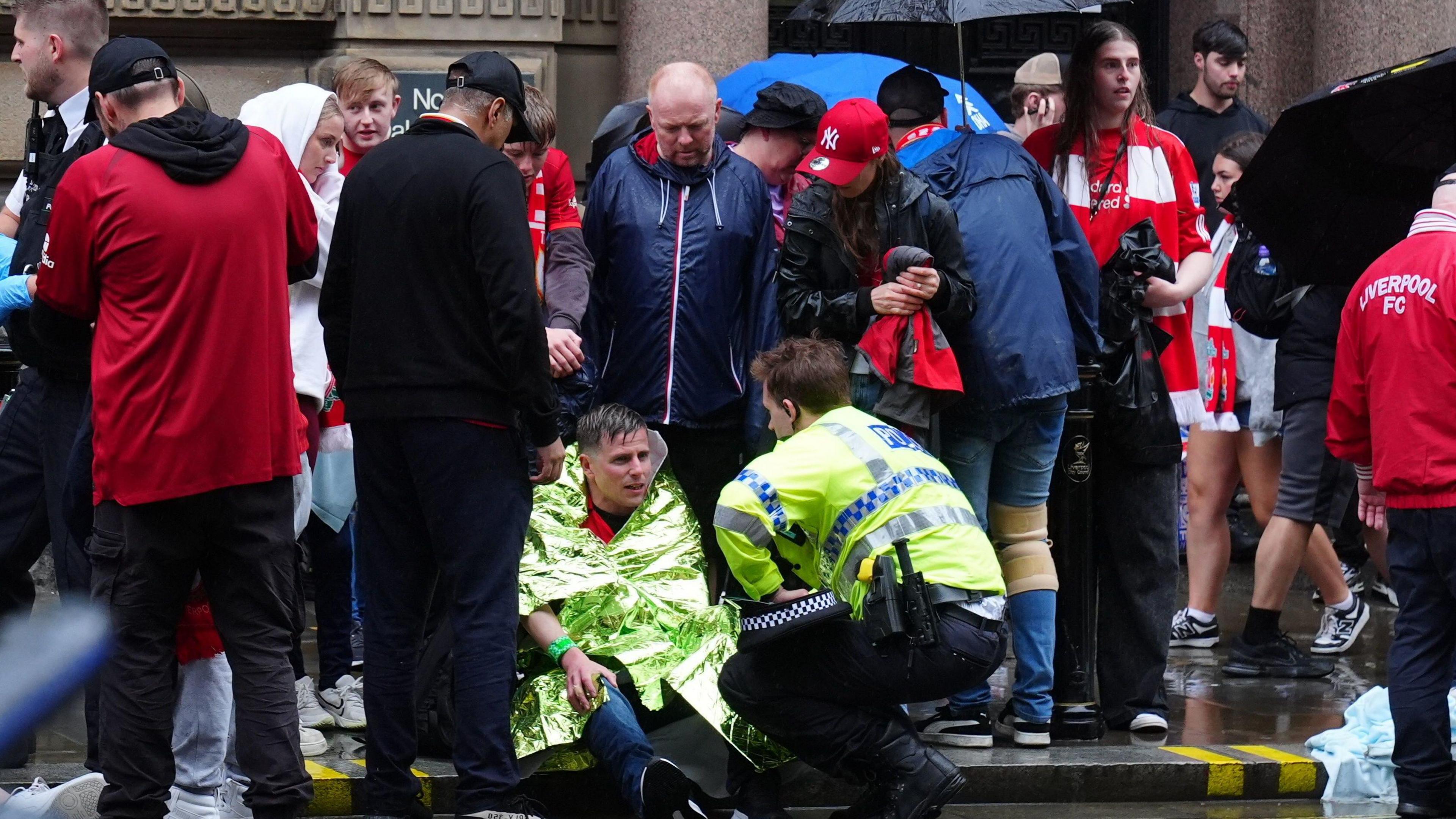 Police officer crouched down talking to man wrapped in emergency service blanket at he sits on the floor. Lots of fans are standing behind them in red, some are wearing coats or holding umbrellas to protect them from the rain