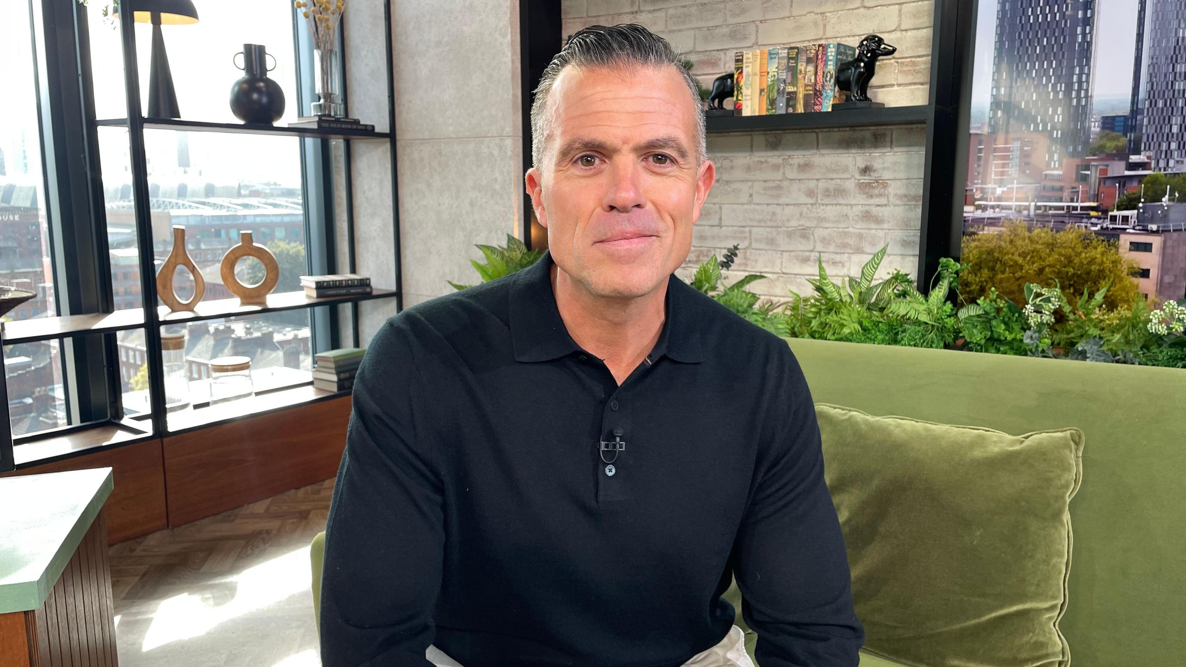 Former detective Marc Cananur in the Morning Live studio, sitting on a green sofa and wearing a dark shirt, next to a window from which Manchester city Centre is visible.