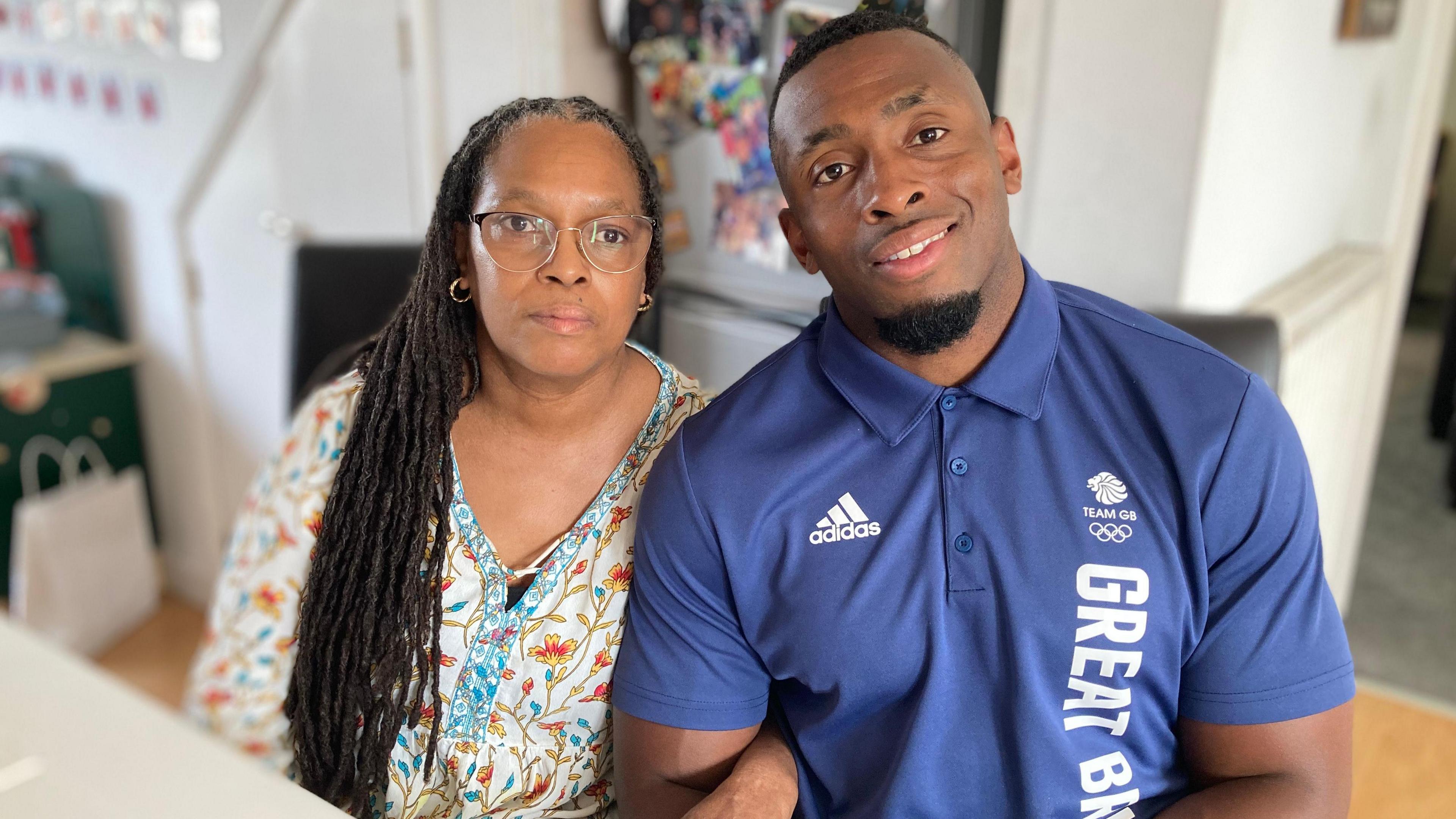 Jacqui Morais and son Joel Fearson sit together with their arms linked. Jacqui is wearing a white shirt with colourful flowers