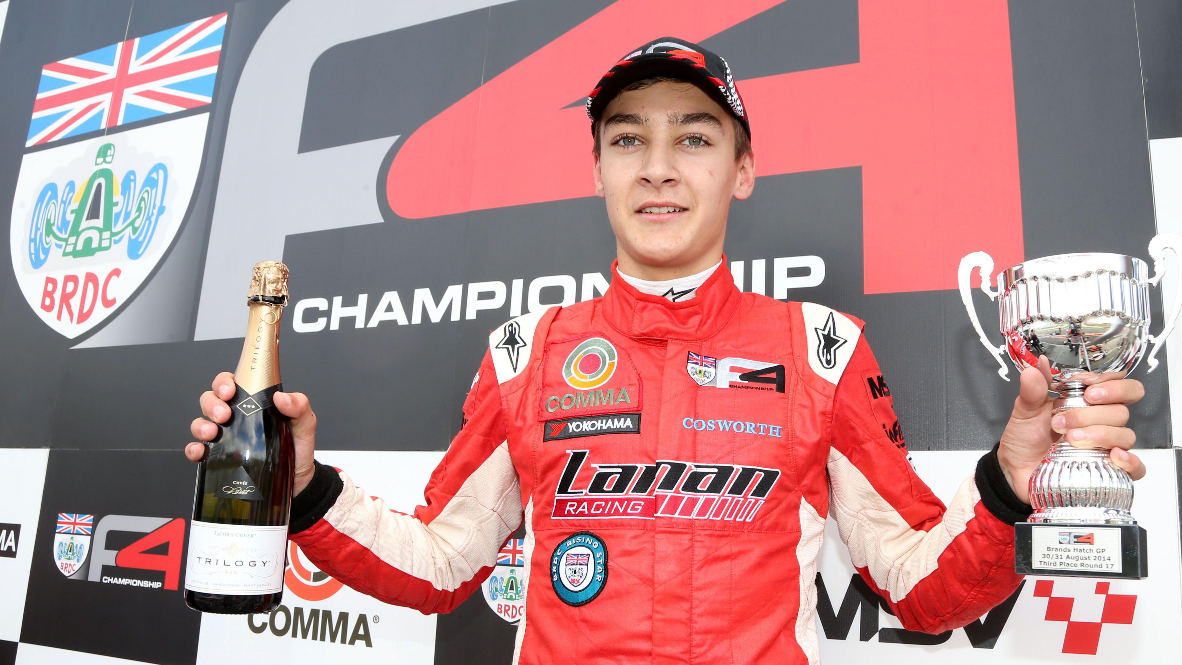 George Russell celebrates on a podium after a Formula 4 victory at Brands Hatch in 2014. He is wearing a red racing suit and his holding a small glass bottle of a drink in his right hand and a trophy in his left hand  