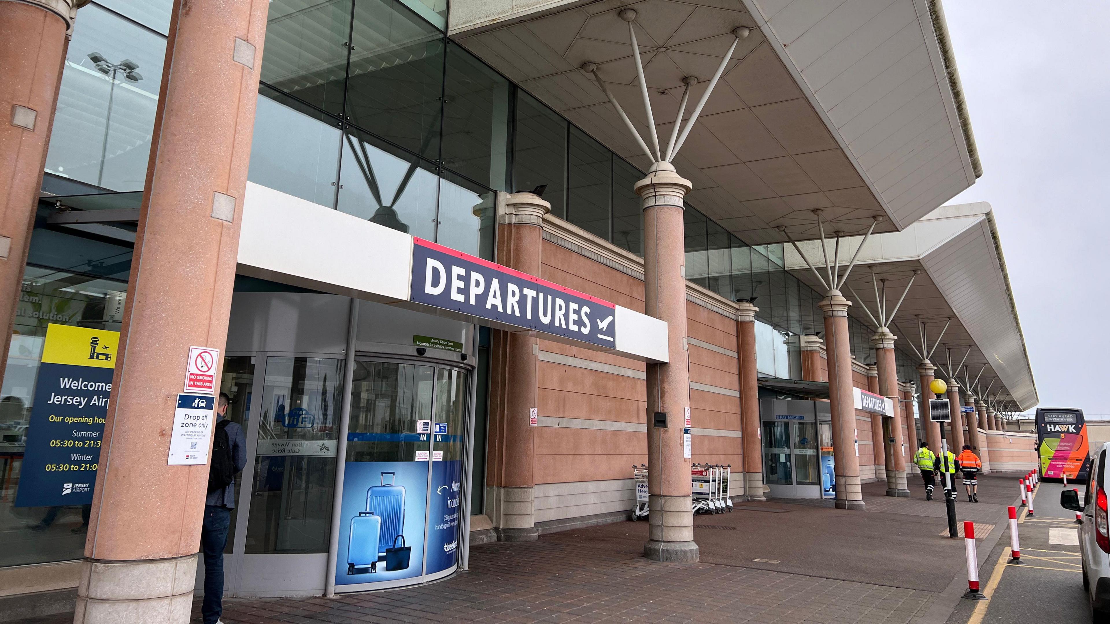 The departures sign is visable outside Jersey Airport as three men in high viz jackets walk away in the background