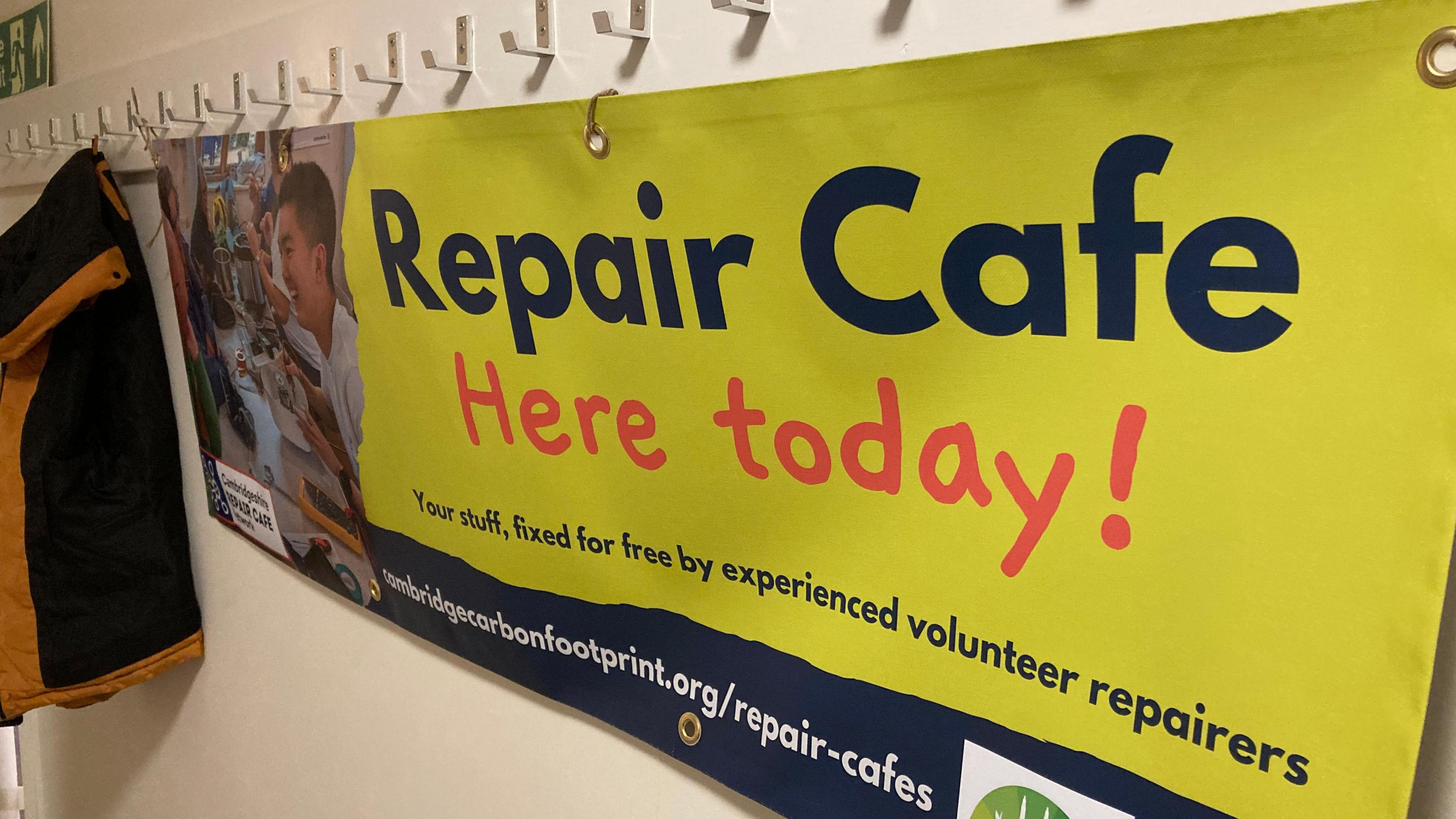 Sign hanging from coat hooks in a corridor, saying "Repair Cafe Here today" on a yellow backing