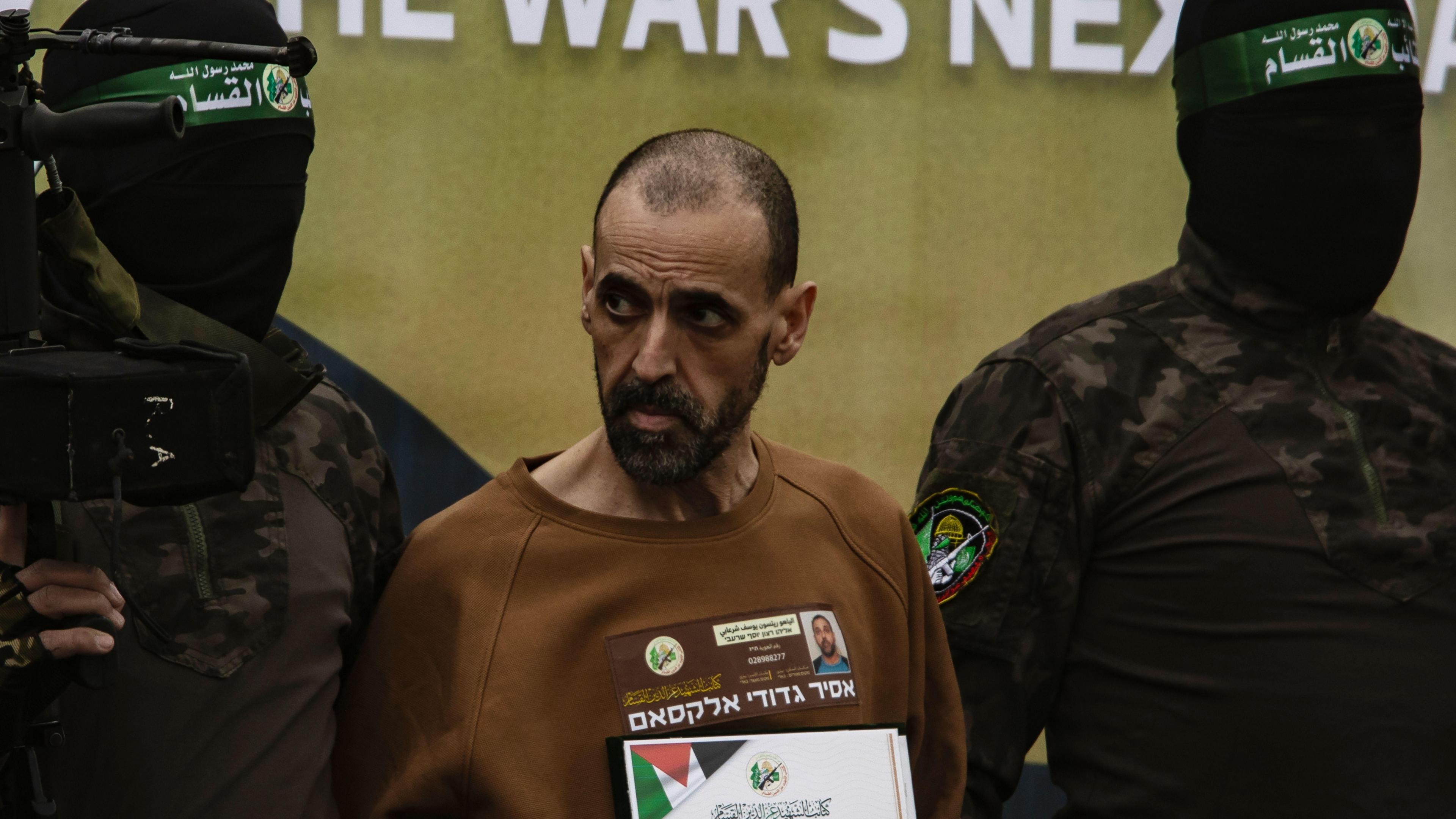 Eli Sharabi flanked by two Palestinian fighters. He is wearing a brown sweatshirt with a banner containing writing and a small picture of a man on. The men either side of him are wearing black face coverings with a green banner on and are wearing long sleeved tops with dark camouflage and a badge on the arm. A gun can be seen in the left hand side of the photo.