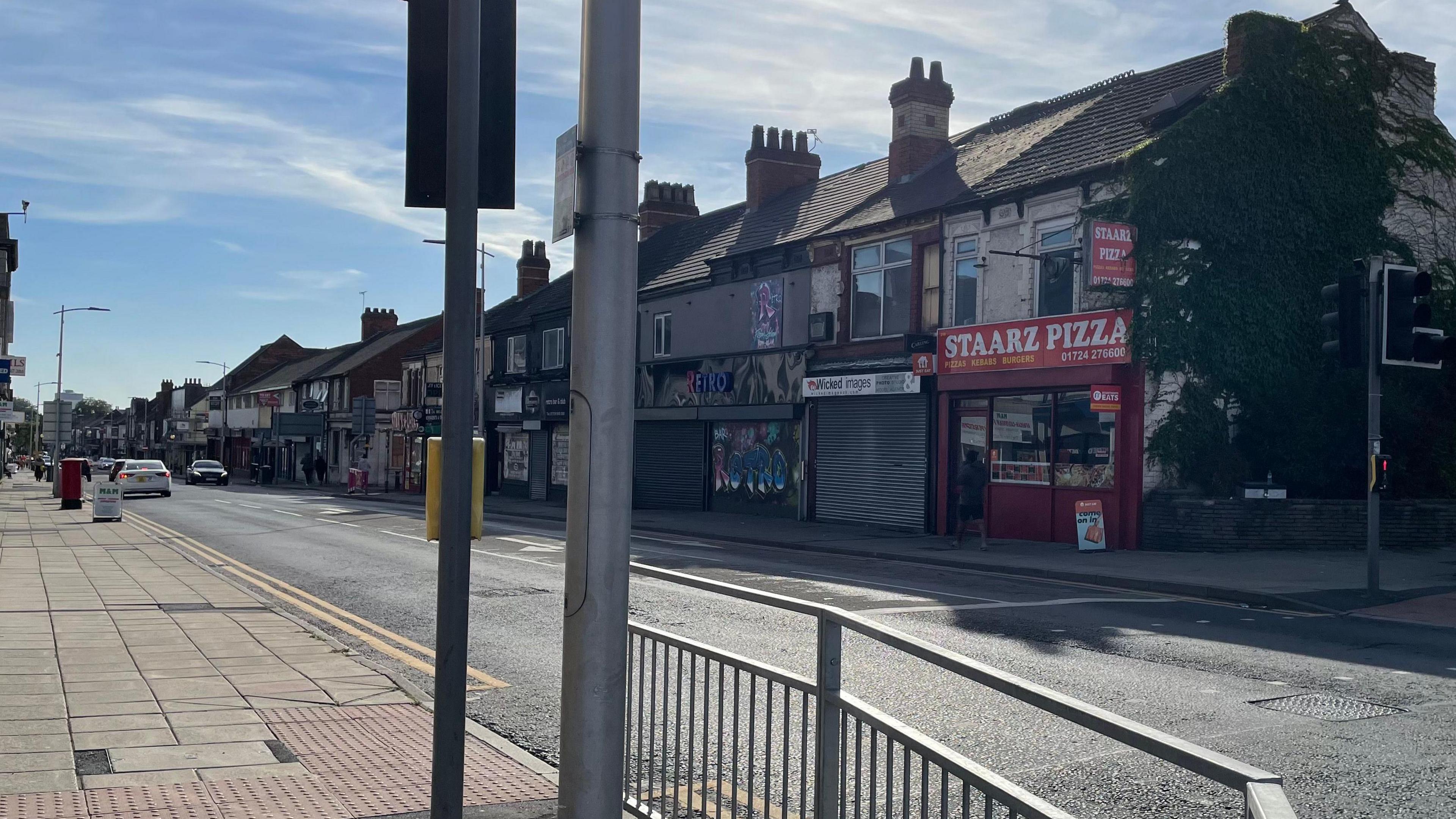 A street with a row of shops and fast-food restaurants. There are traffic lights and a metal fence.