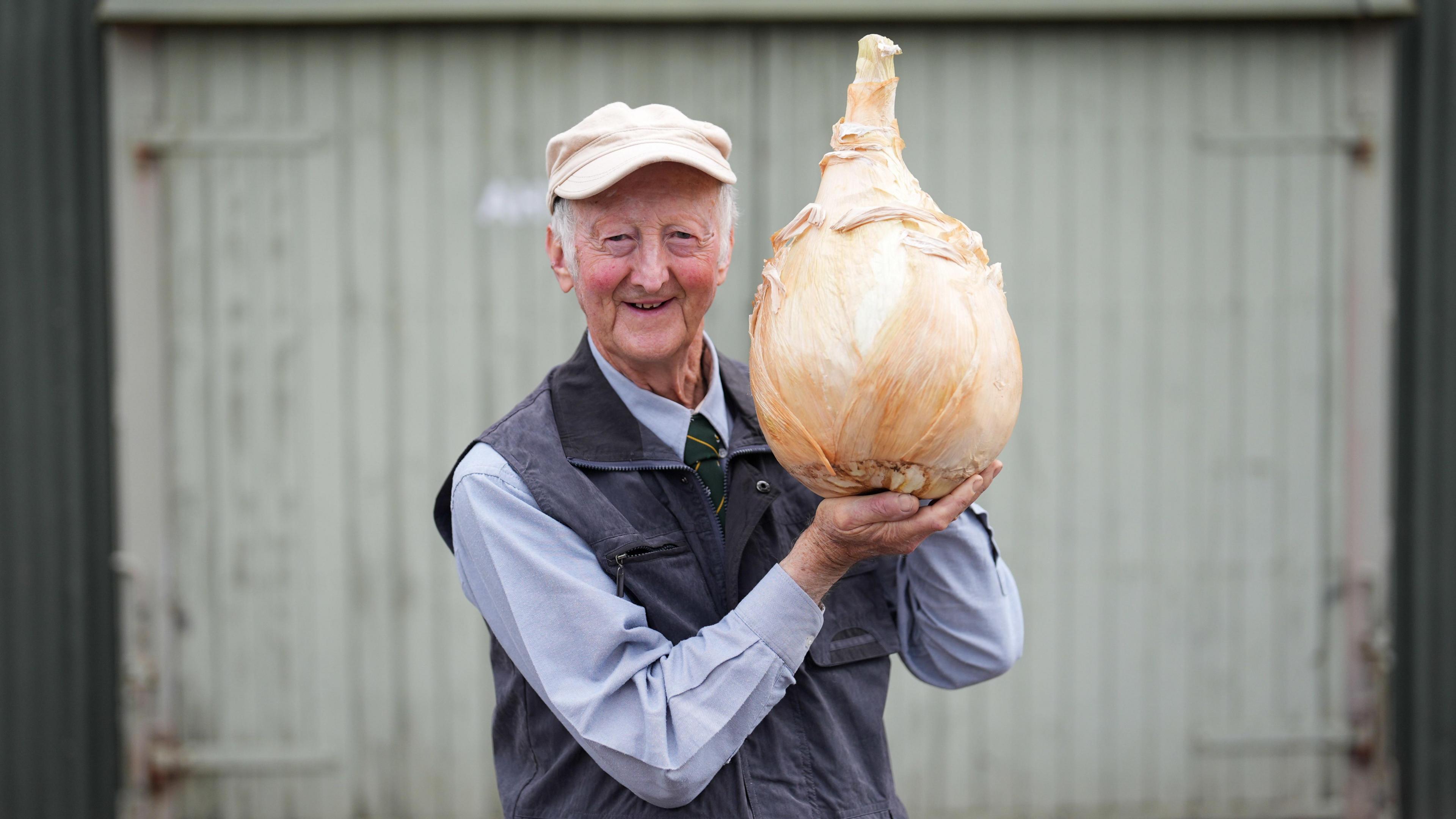 A man in a pale blue shirt and grey gilet holds up a giant onion.