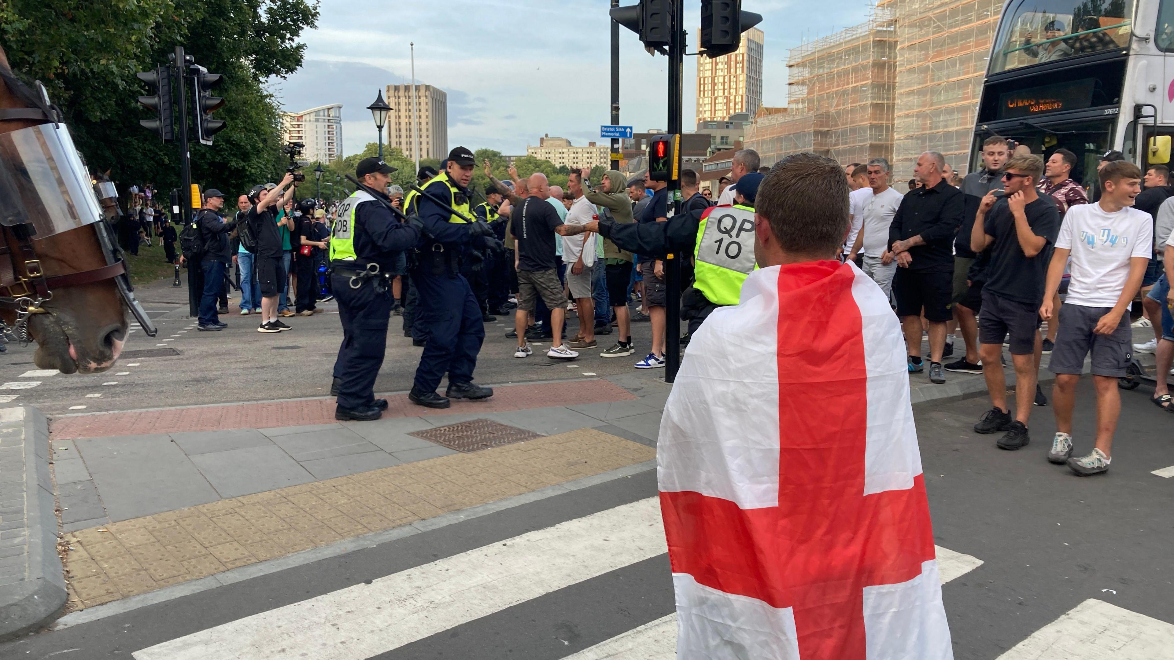 A man draped in an England flag looks on at protestors in the street