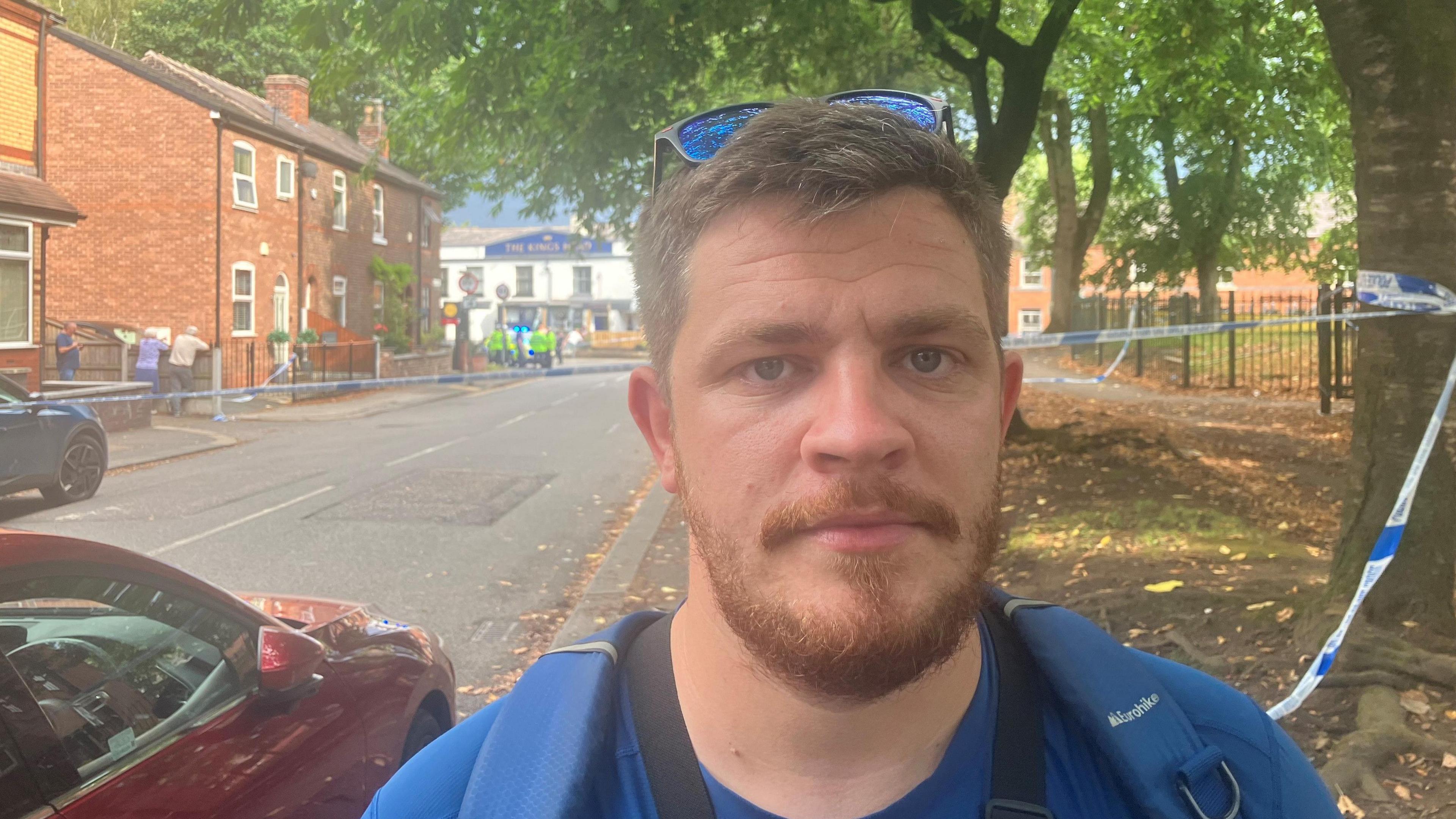 A man with a gingerish beard and sunglasses on his head stands wearing a backpack next to a road. Behind him the road has been blocked off with police tape.