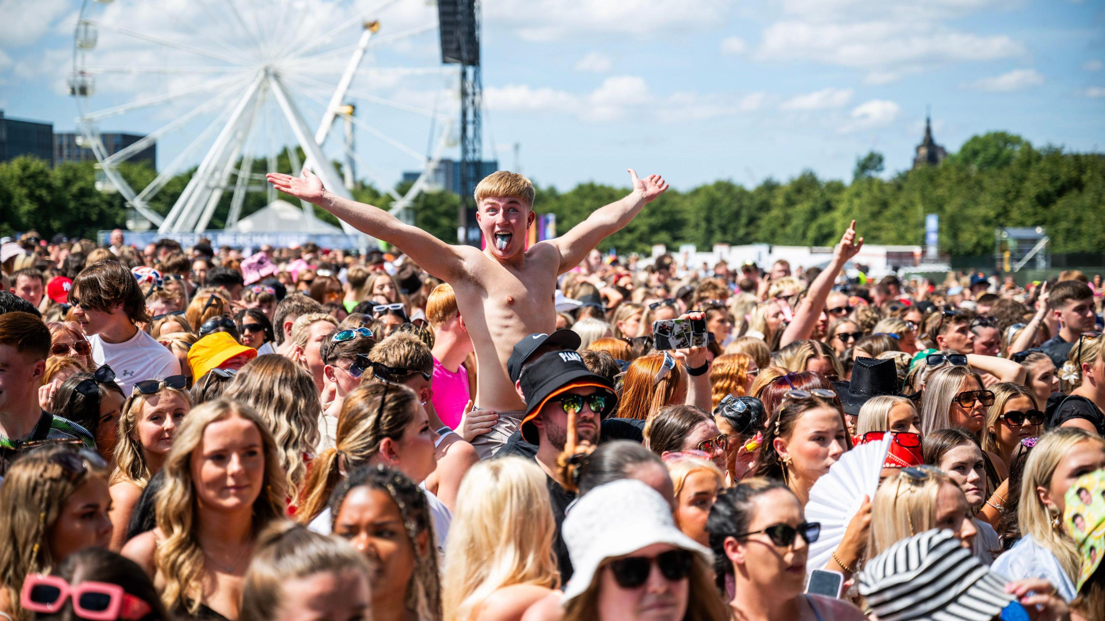 A large crowd at a music festival on a sunny day. Among the crowd are a shirtless fan, who sticks his tongue out while stretching his arms out wide as he sits on a friend's shoulders.  A large ferris wheel can be seen in the background. 