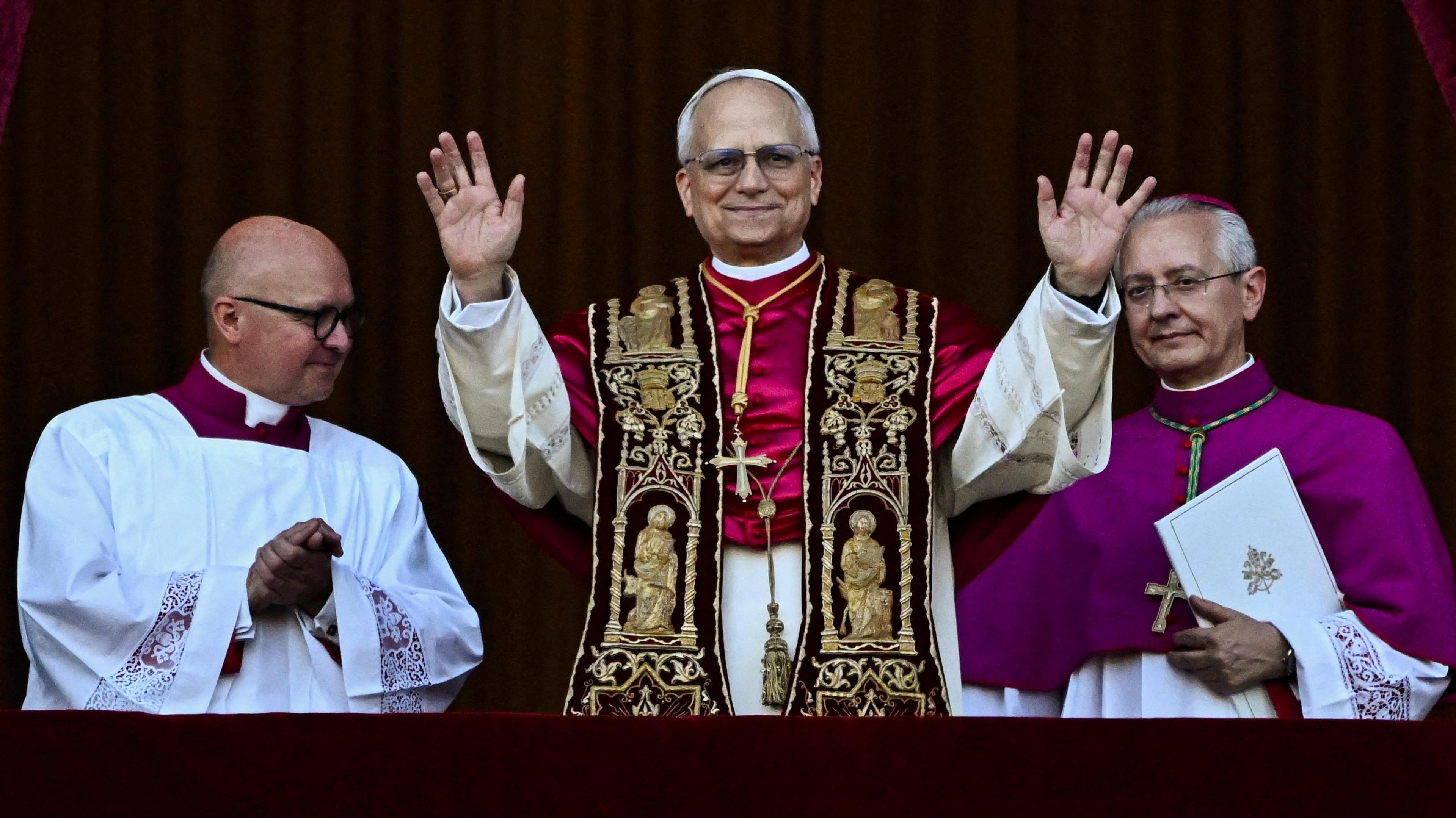Pope Leo XIV stands on the balcony before the crowd in St Peter's Square, holding his hands in the air alongside two Vatican officials