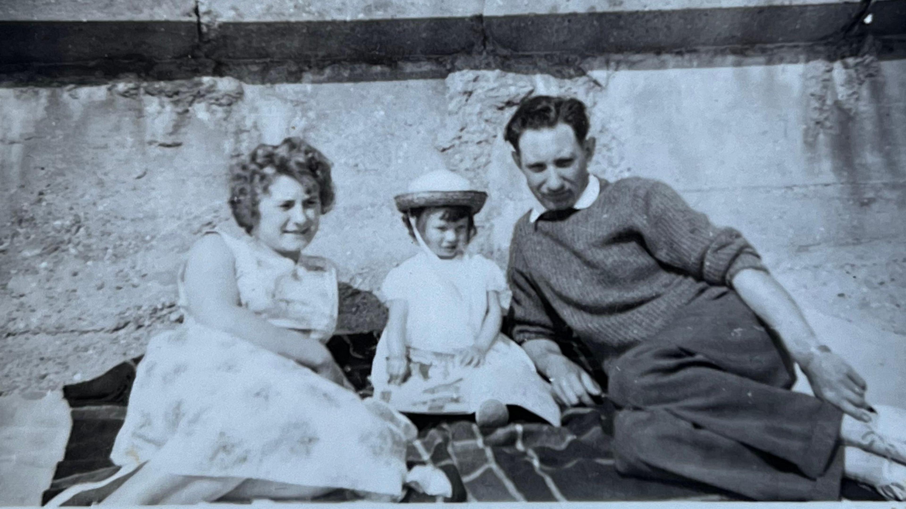 An old black and white photo of a woman and a man lying on a tartan picnic blanket with their young daughter sat between them in a little bonnet.