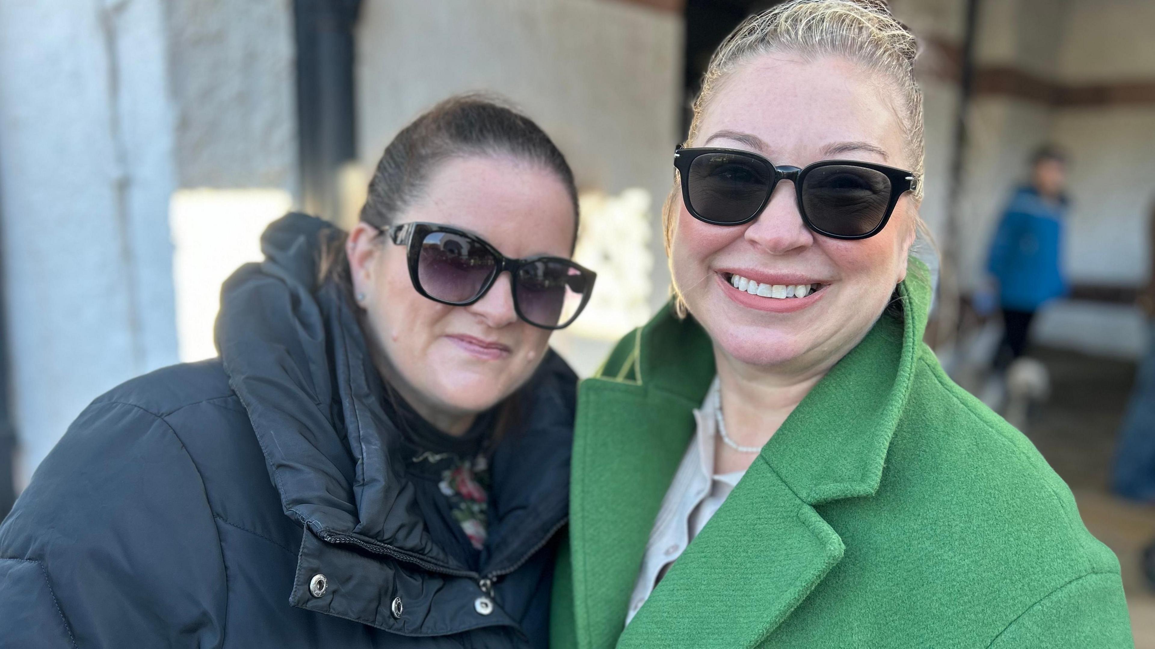 Emma Welton and Andrea Asbridge-Heard, who are both wearing sunglasses. One is brunette and is wearing a black down jacket, and the other is blonde and wearing a green coat. 