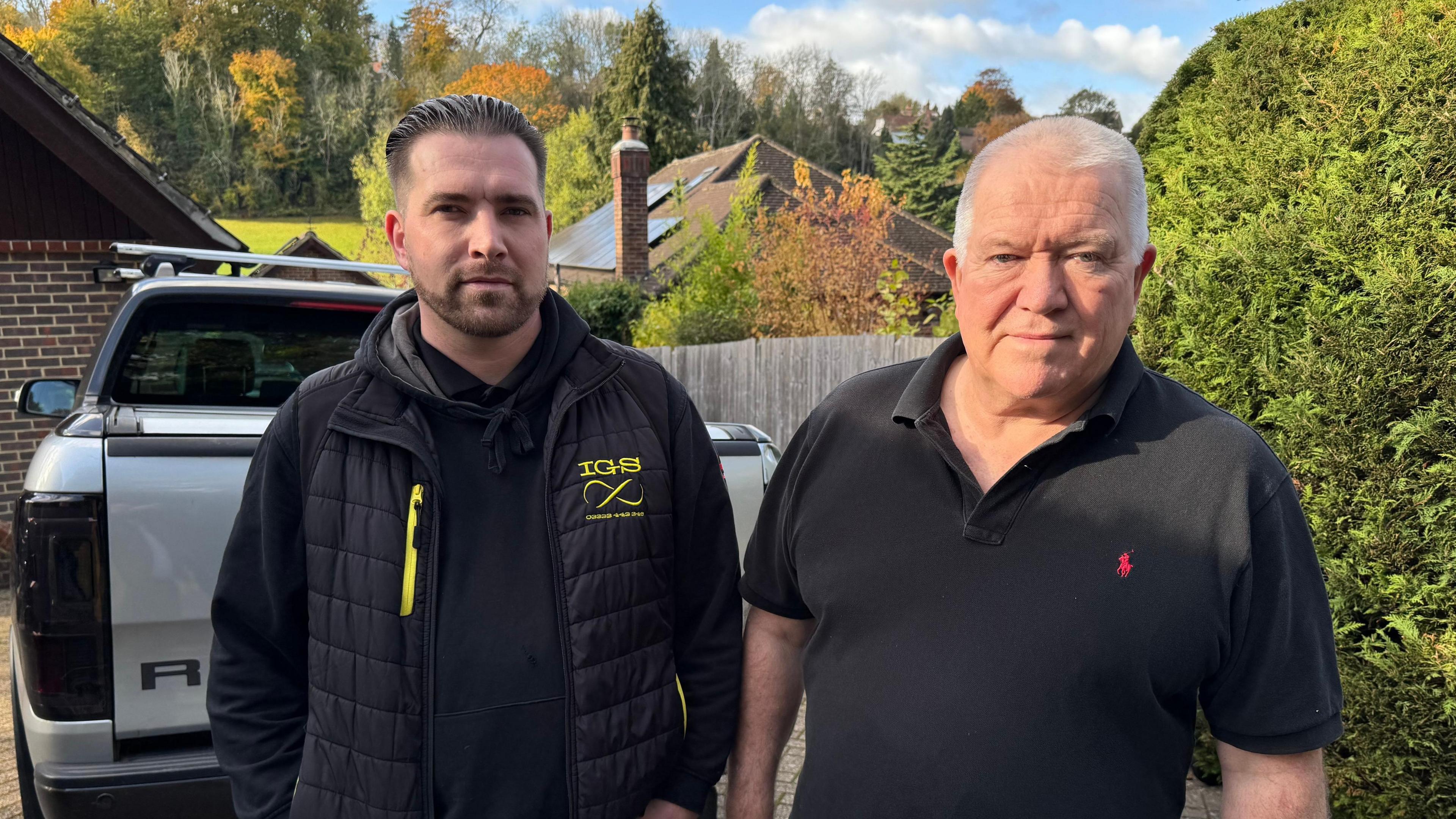Steve Piller, with black hair and a beard and dressed in black, stands in front of his truck and next to his dad, Simon, who has short white hair and is wearing a black t-shirt.