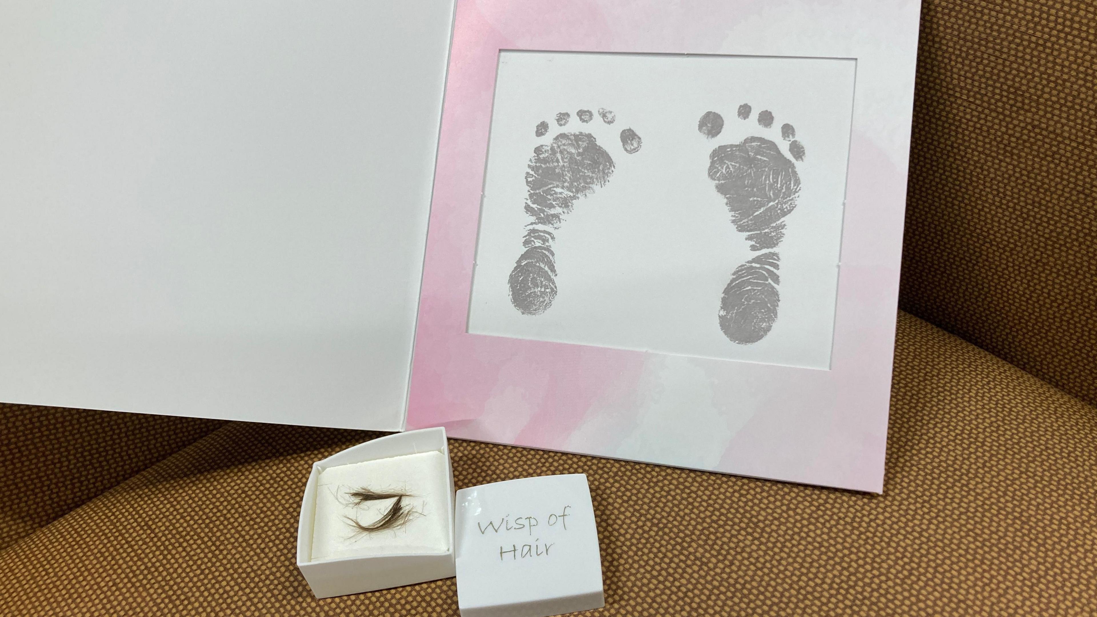 A picture of two grey small baby footprints are seen in a card pink-framed card.  A box with two small pieces of hair is open and on the lid read 'wisp of hair'.
