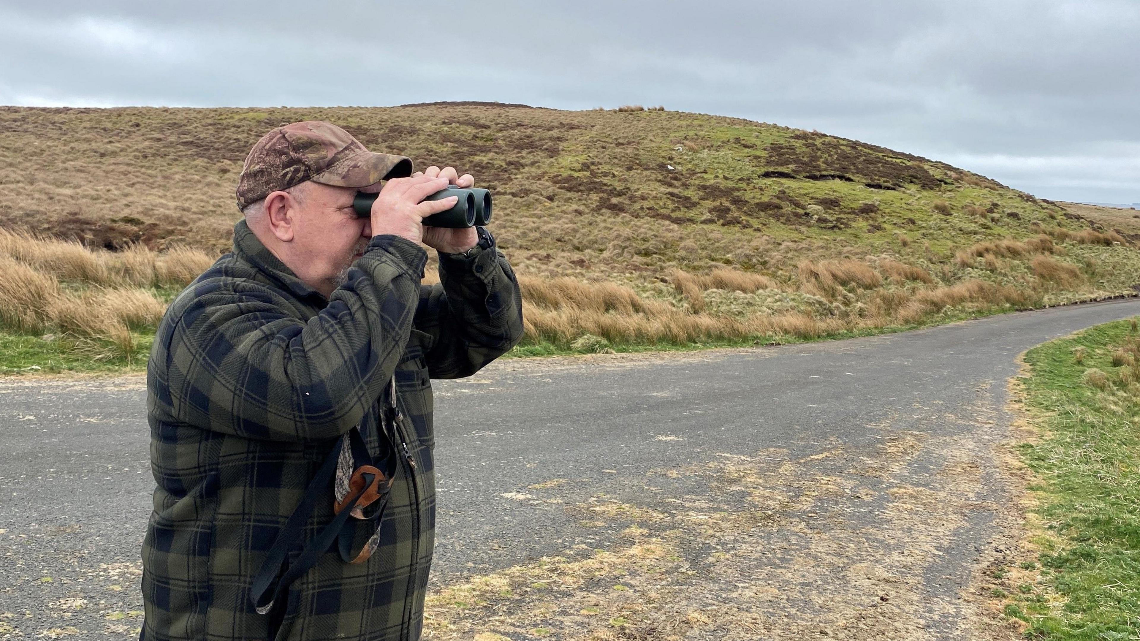 Ian Glendinning is holding binoculars to his eyes and staring across an empty moorland landscape. He is wearing a green and black check jacket and brown peaked cap.