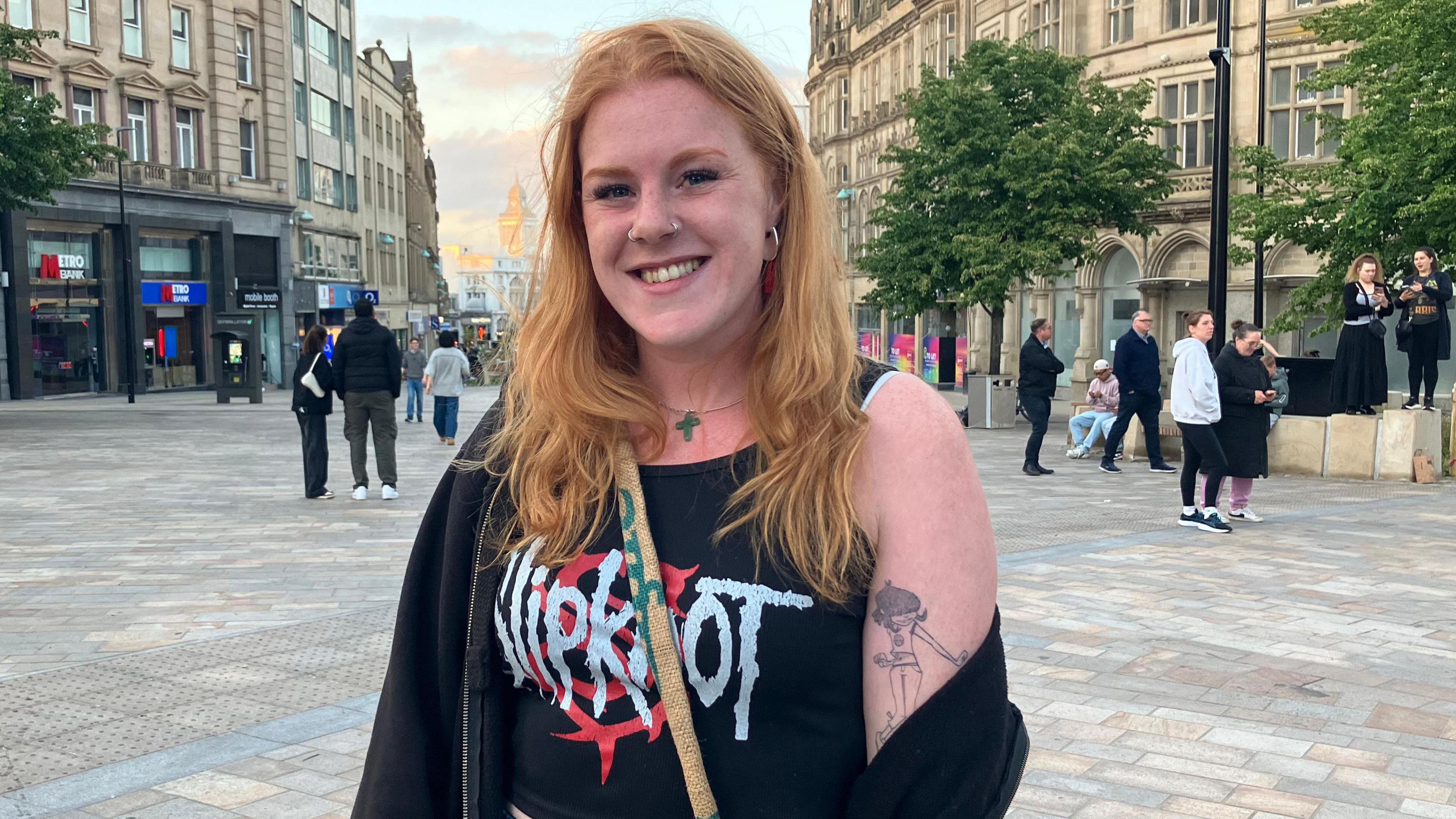 A woman stands in a square. She has ginger hair that reaches past her shoulders and is wearing a black t-shirt and black cardigan. A tattoo is on her left upper arm and she is wearing a bag.