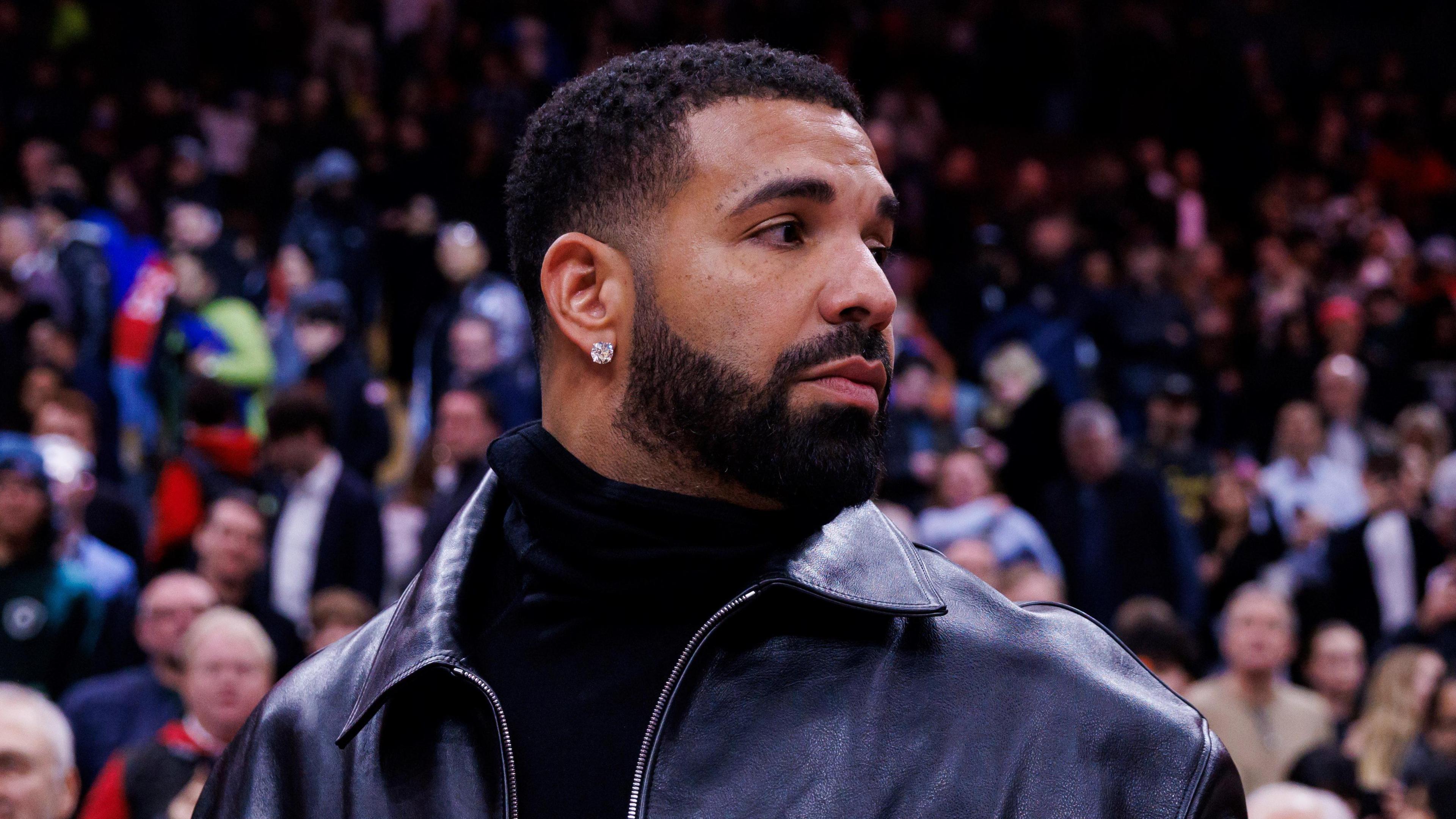 Drake pictured at a basketball game in January. The rapper looks over his left shoulder, wearing a black leather jacket over a black turtle-neck jumper. He has a diamond earring in his right ear. Behind him is a crowd of basketball fans. 