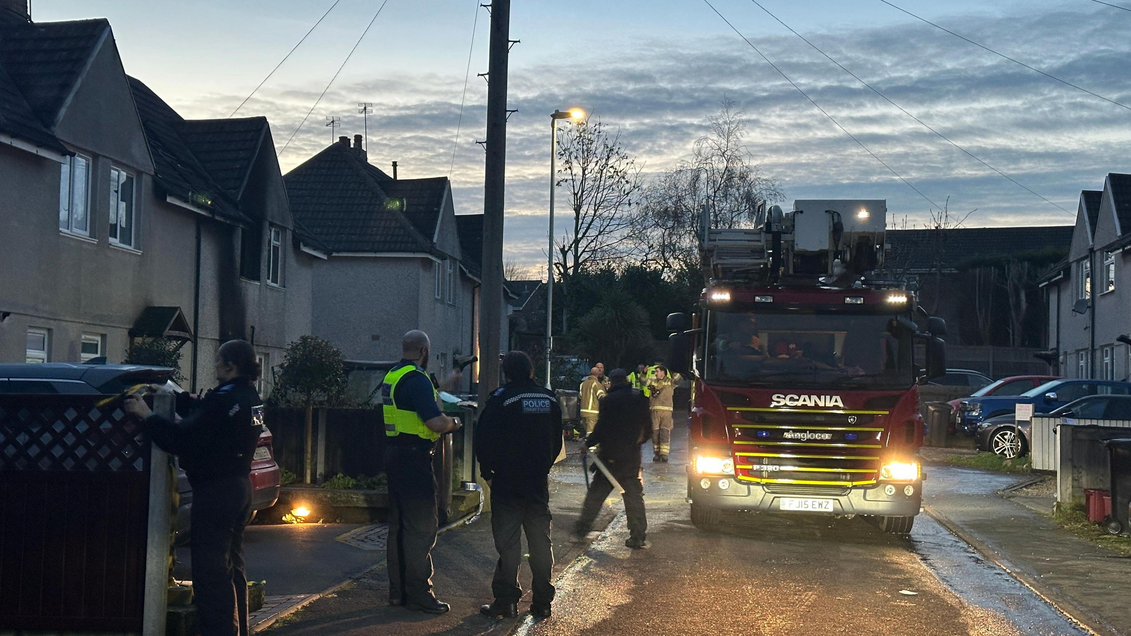 A fire engine is parked in a road outside a fire damaged house. Police officers and firefighters can be seen standing on the pavement along the row of houses.