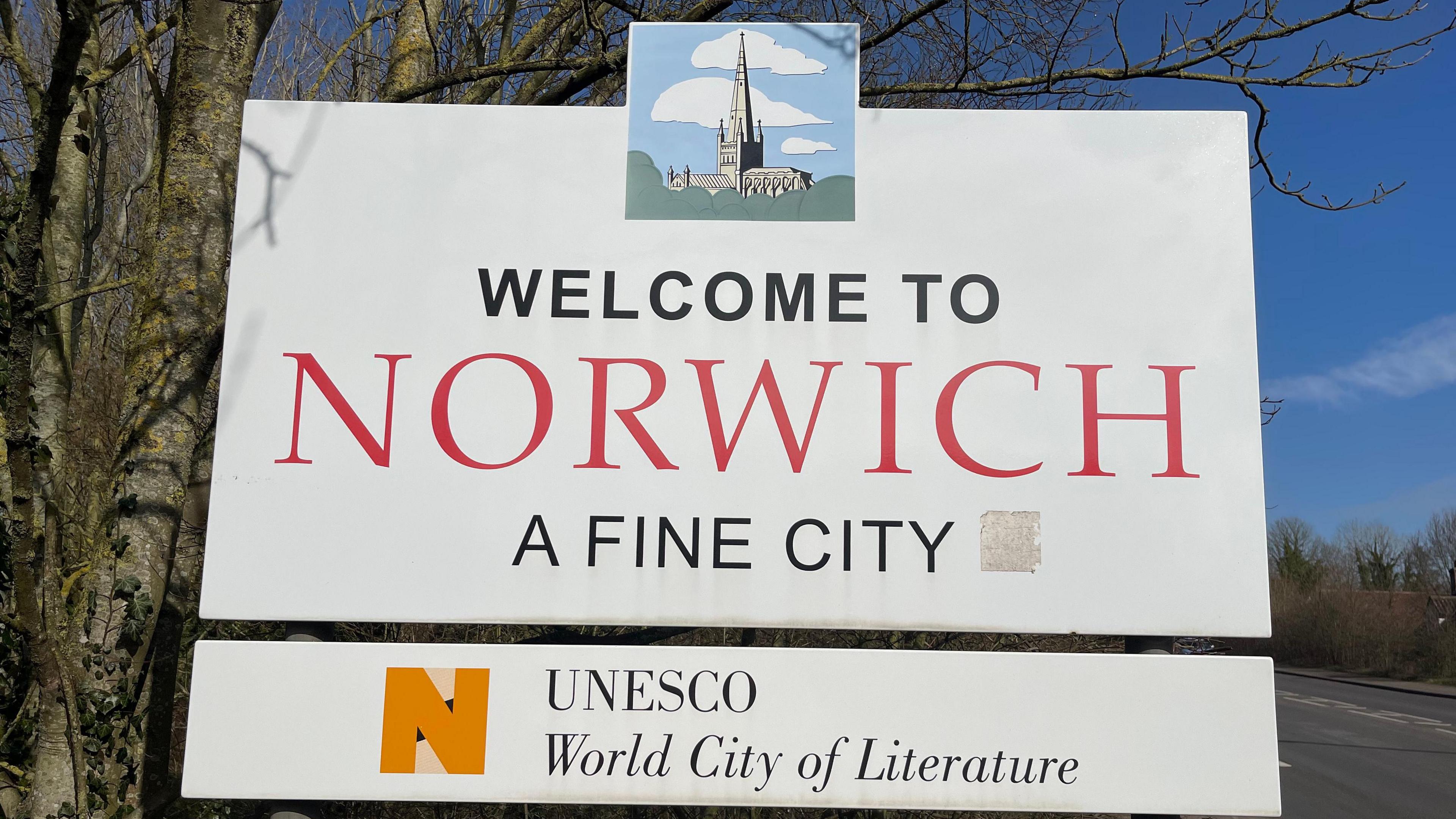 A white Norwich sign welcoming people to the city. The sign says WELCOME TO NORWICH, A FINE CITY. UNESCO World City of Literature.