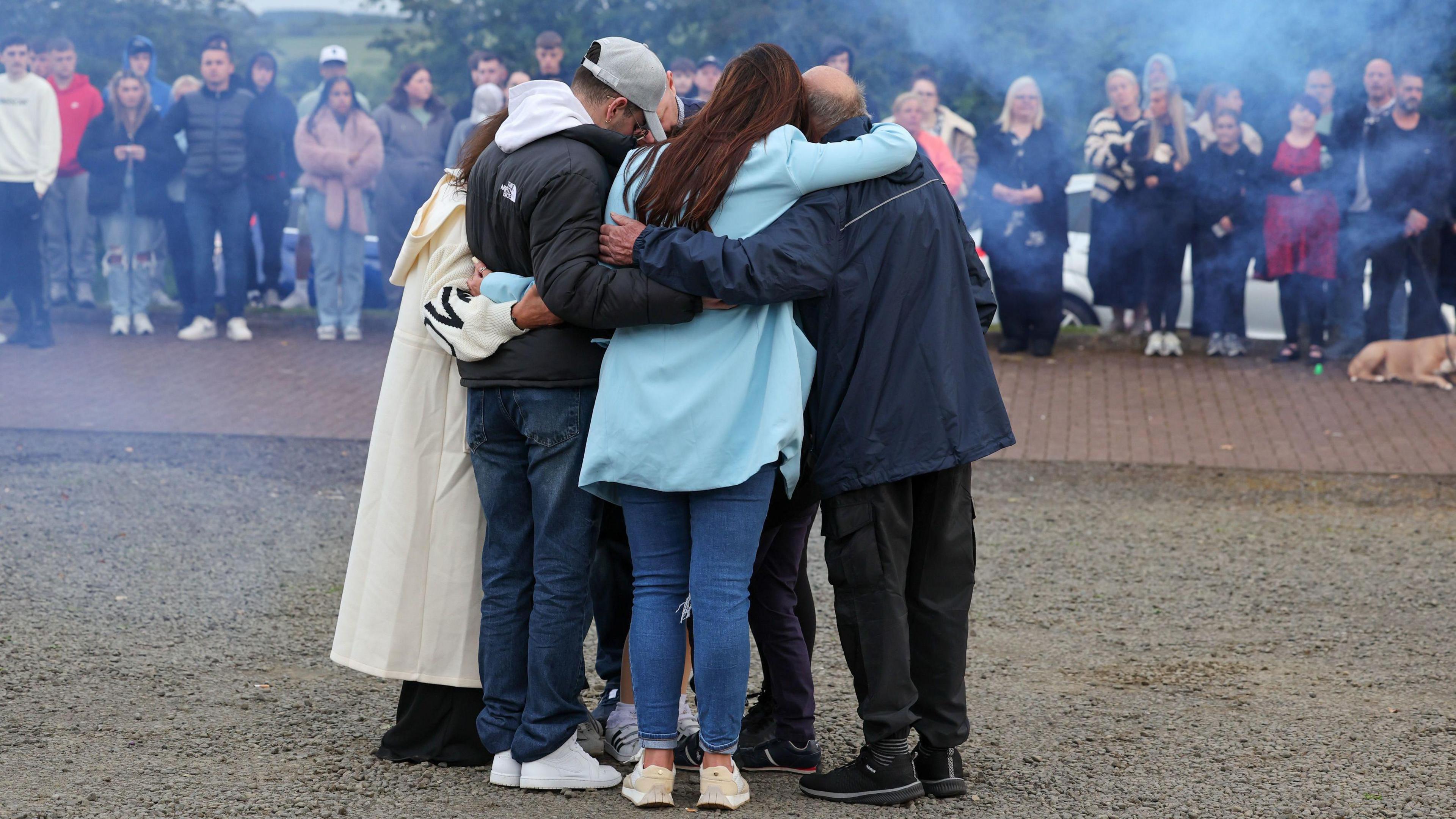 Mourners line the road behind a group of Cole Cooper's family members huddled together in an embrace at his vigil. Blue smoke cannisters have been released around them to indicate his favourite colour.