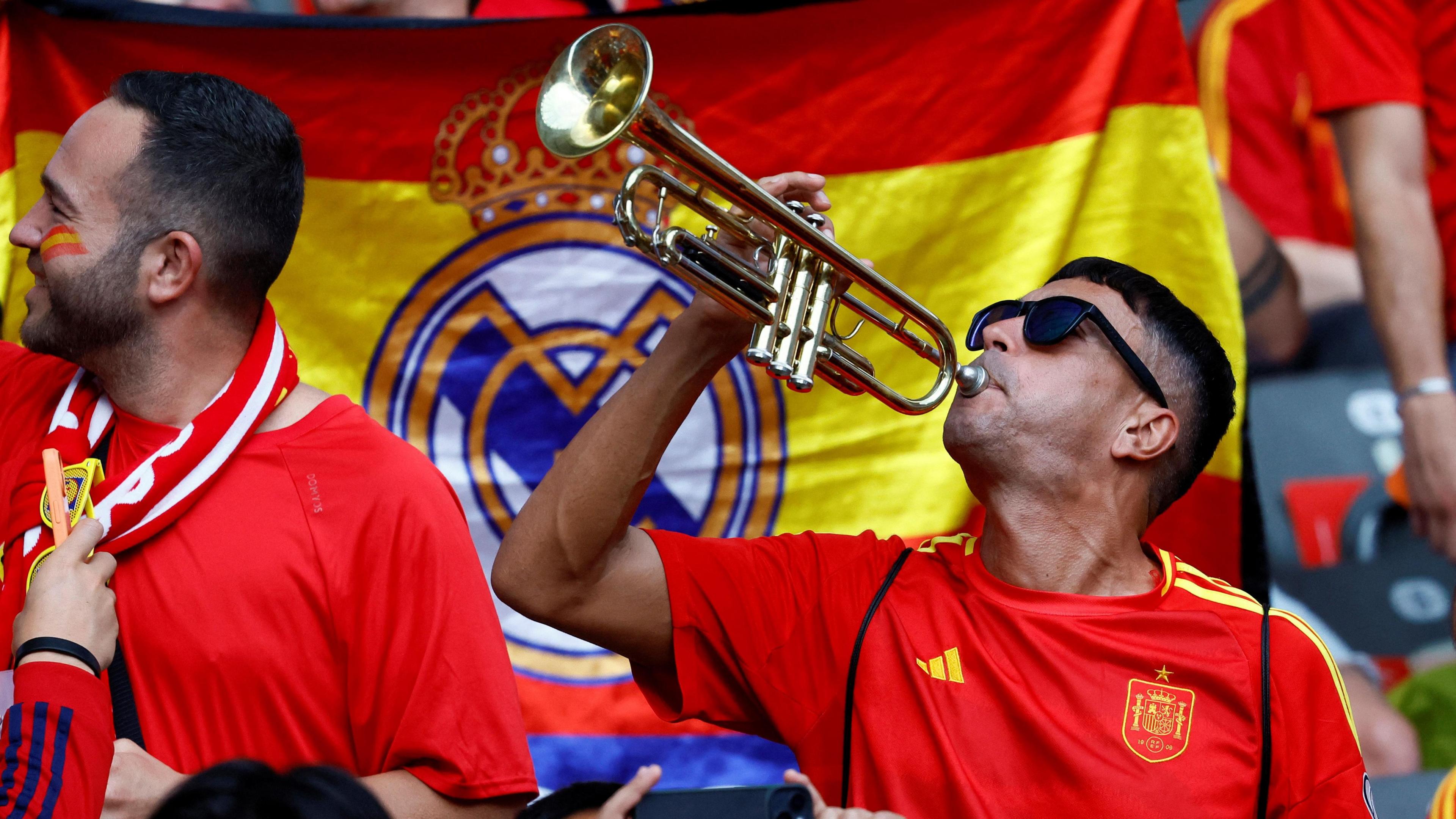 A Spain supporter plays a brass instrument in front of a Spanish flag in a crowd of fans