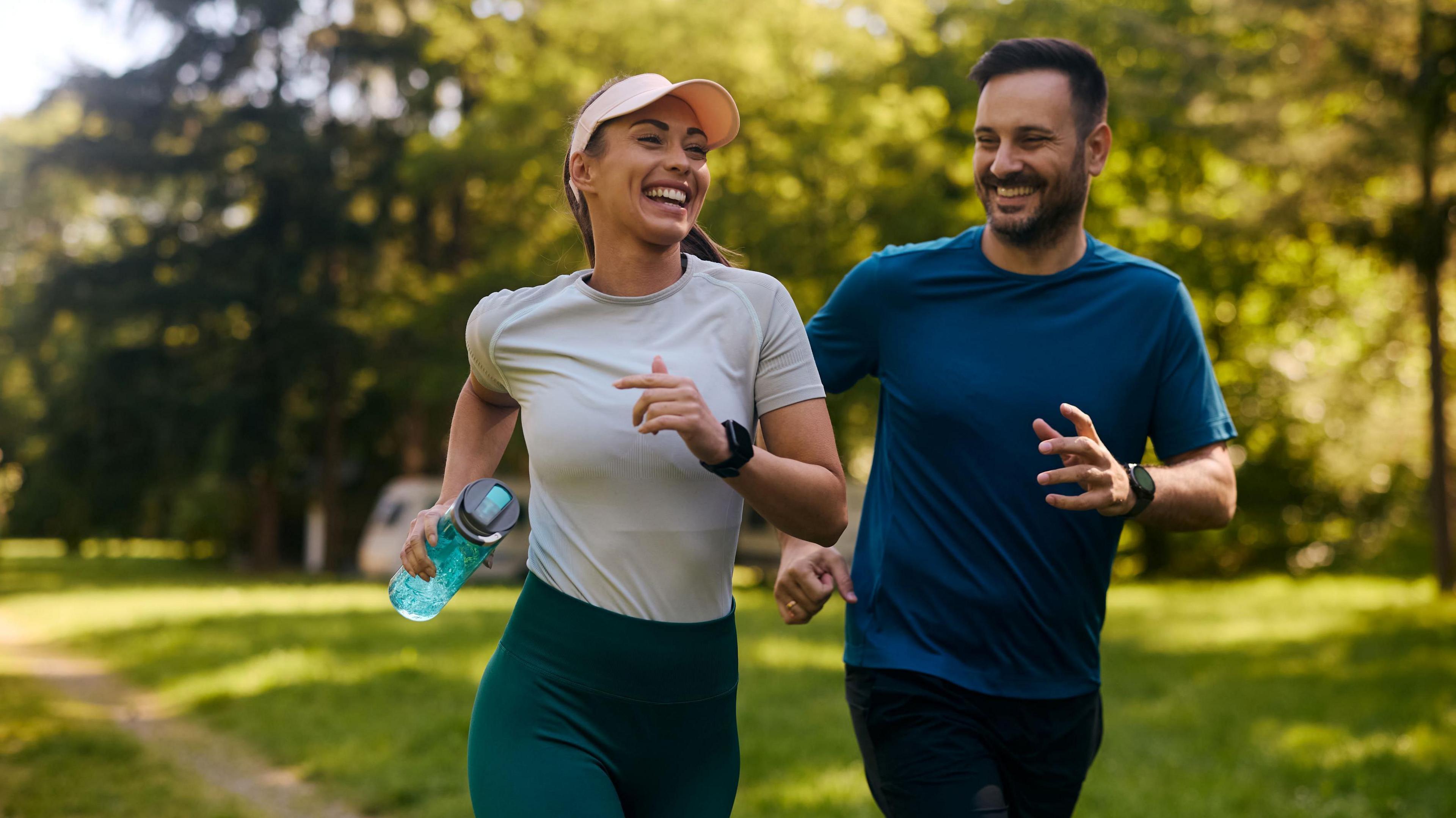 Two people - a man and a woman - smiling as they jog through a park on a sunny day. The woman is wearing a light coloured top, a pink cap, dark green running leggings and is holding a blue plastic drinks bottle, while the man is wearing a dark blue T-shirt and black bottoms. They are both smiling.