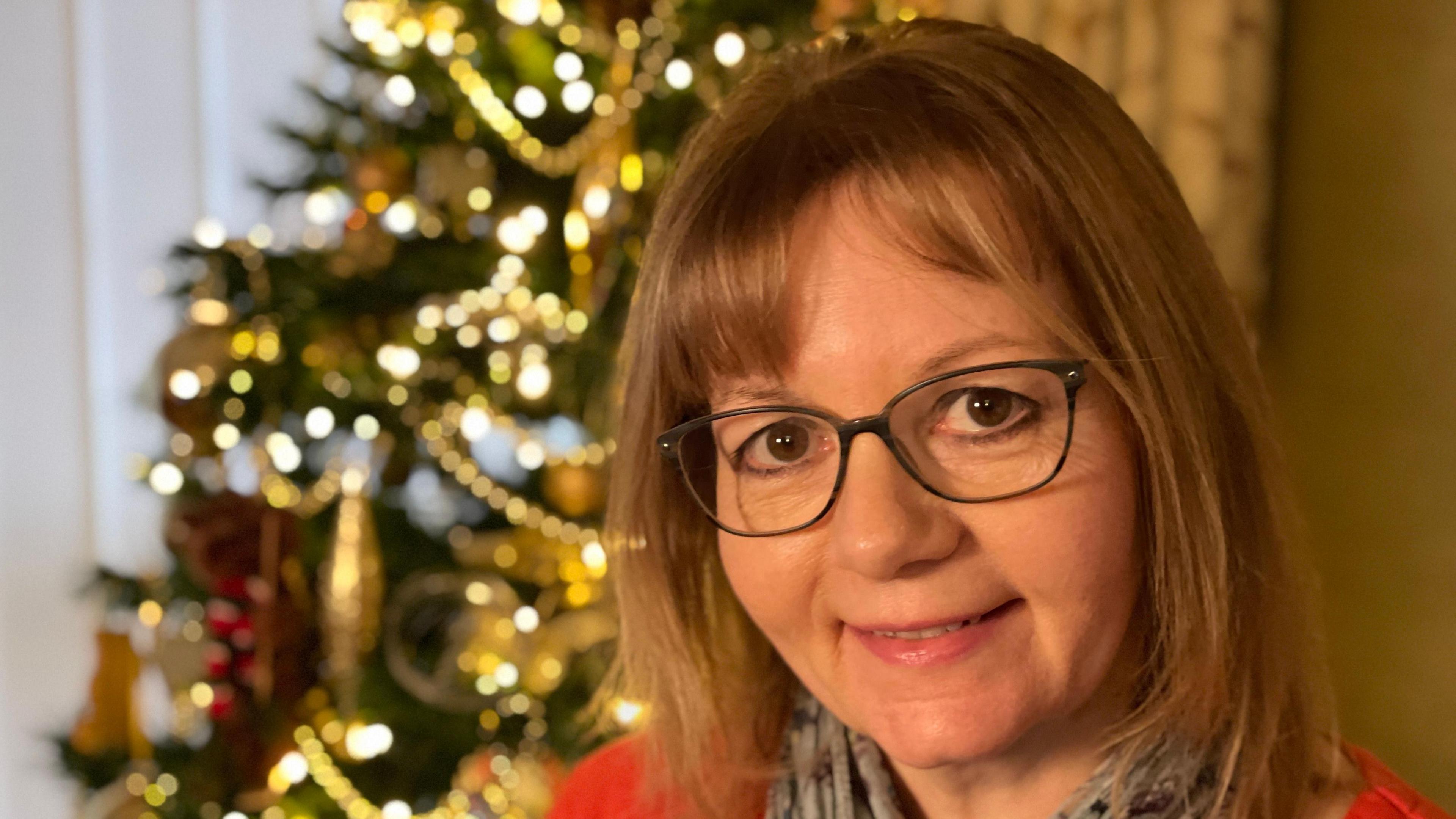 Ms Bretton smiles at the camera in front of a Christmas tree. She has red hair in the style of a bob and has black glasses. She wears a red jumper with a grey scarf. 