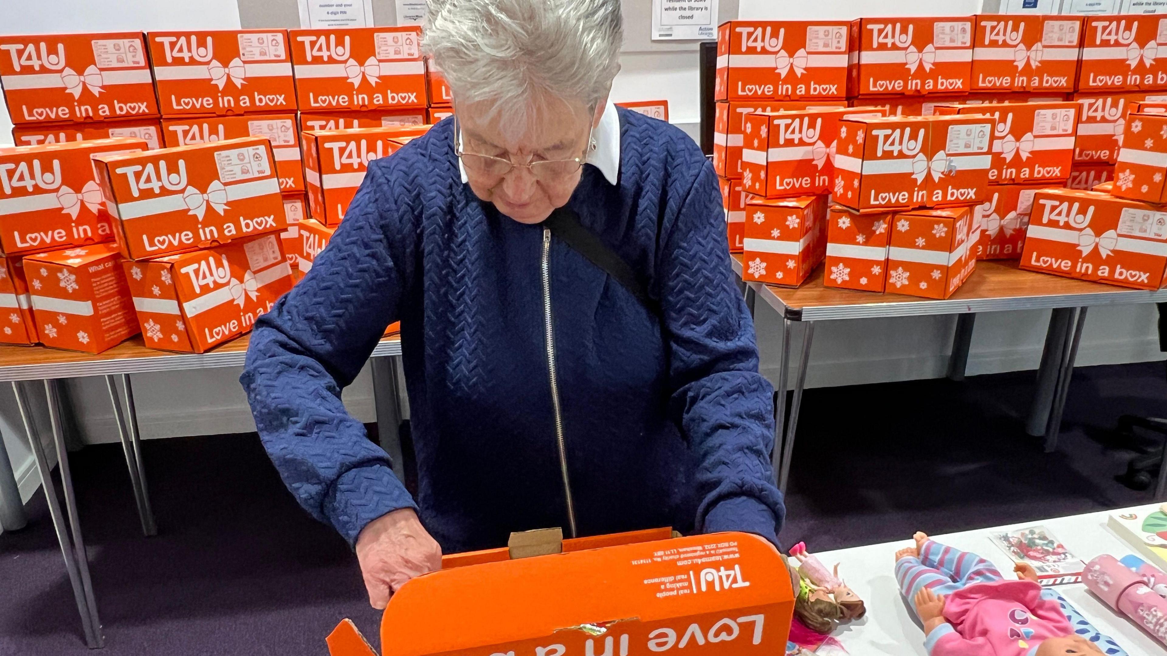 Maureen Summerhayes looking at a shoebox with love in a box written on it.  as she packs it with gits. Maureen is wearing a navy zipped cardigan with a white blouse