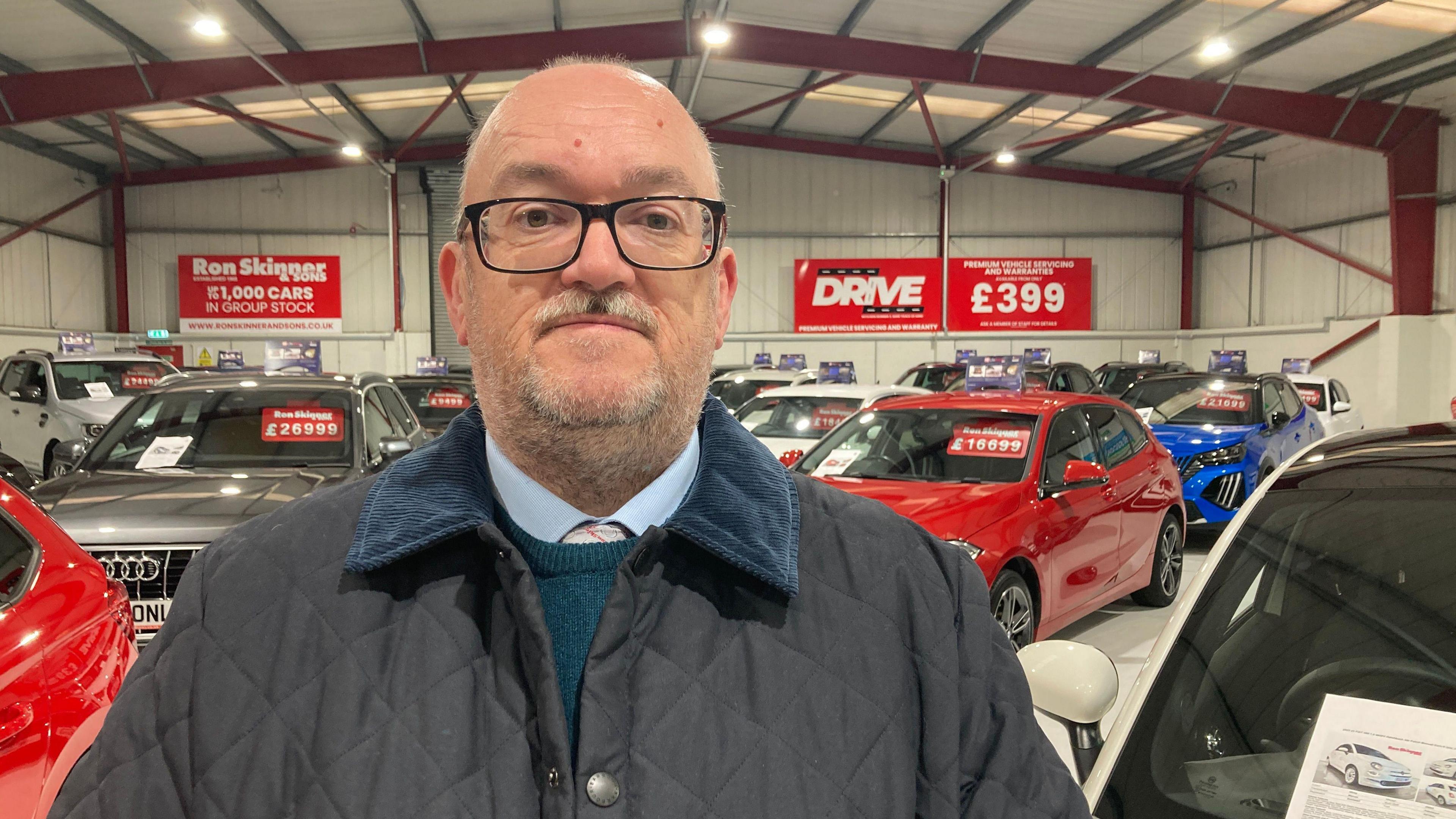 Philip Skinner in a dark blue jacket stands in a car showroom with different colour cars in the background with price tags on and Ron Skinner signs on the wall.