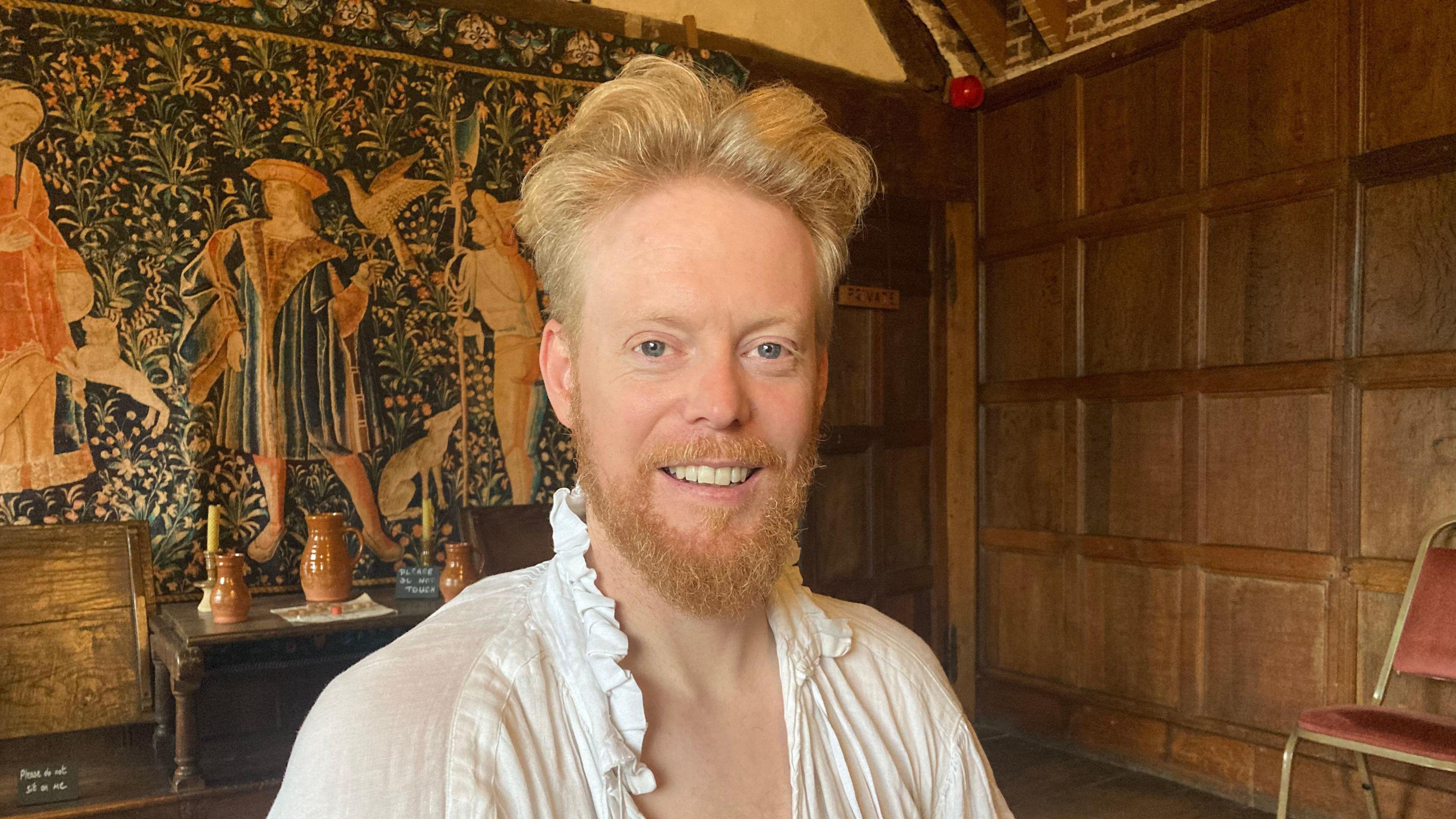 Phil Downing pictured in the manor house with a green and gold tapestry behind him. He has blonde hair and is wearing a 16th Century style night shirt.