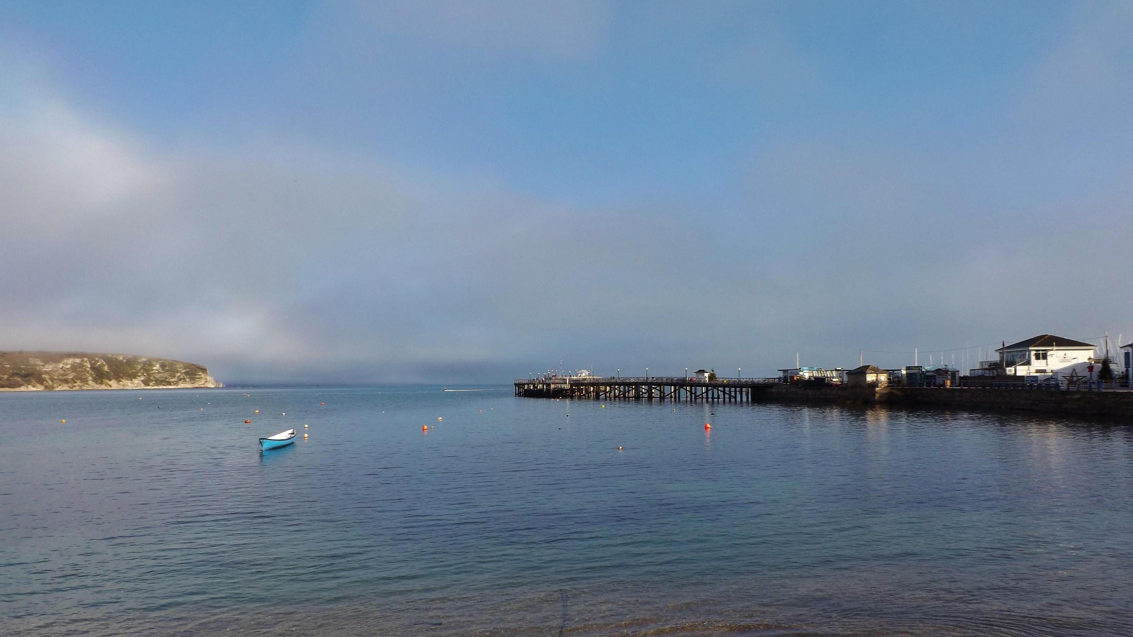 A fairly sunny day overlooking a body of water. A pier runs down the right-hand side with a canoe-like boat floating on the water on the other side.