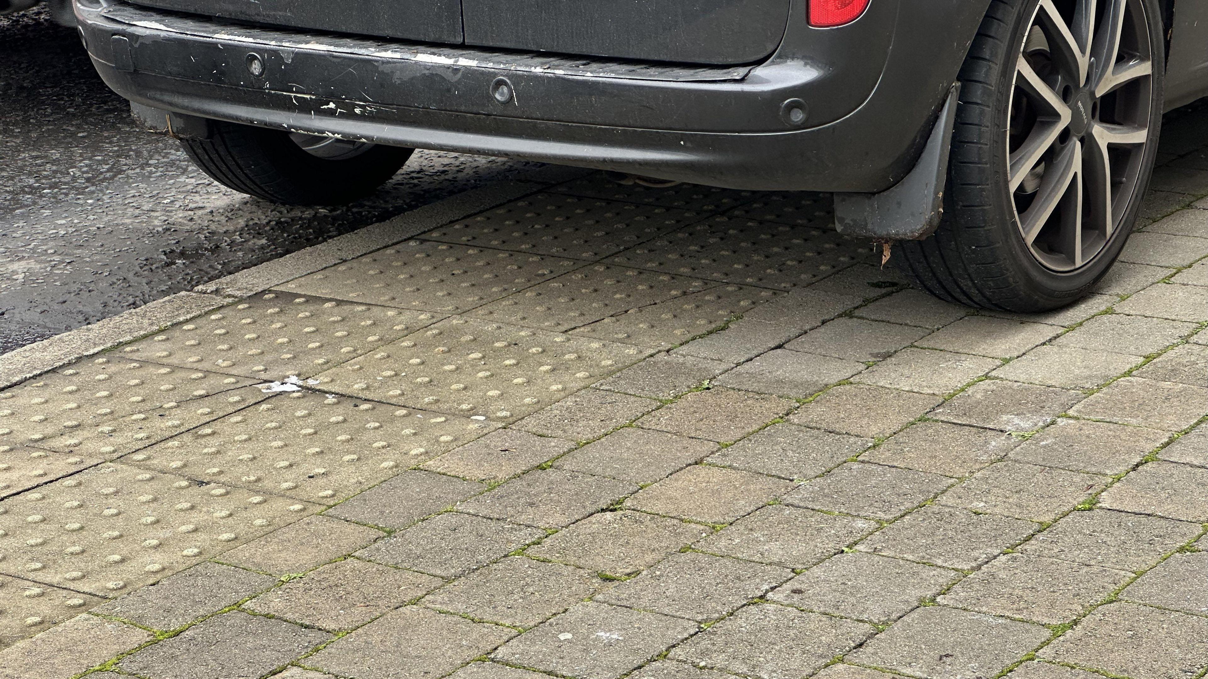 The wheels of a black van are photographed parked on the pavement which includes tactile pavings at a crossing on Main Street. Most of the vehicle is on the path with only its left wheels on the road.