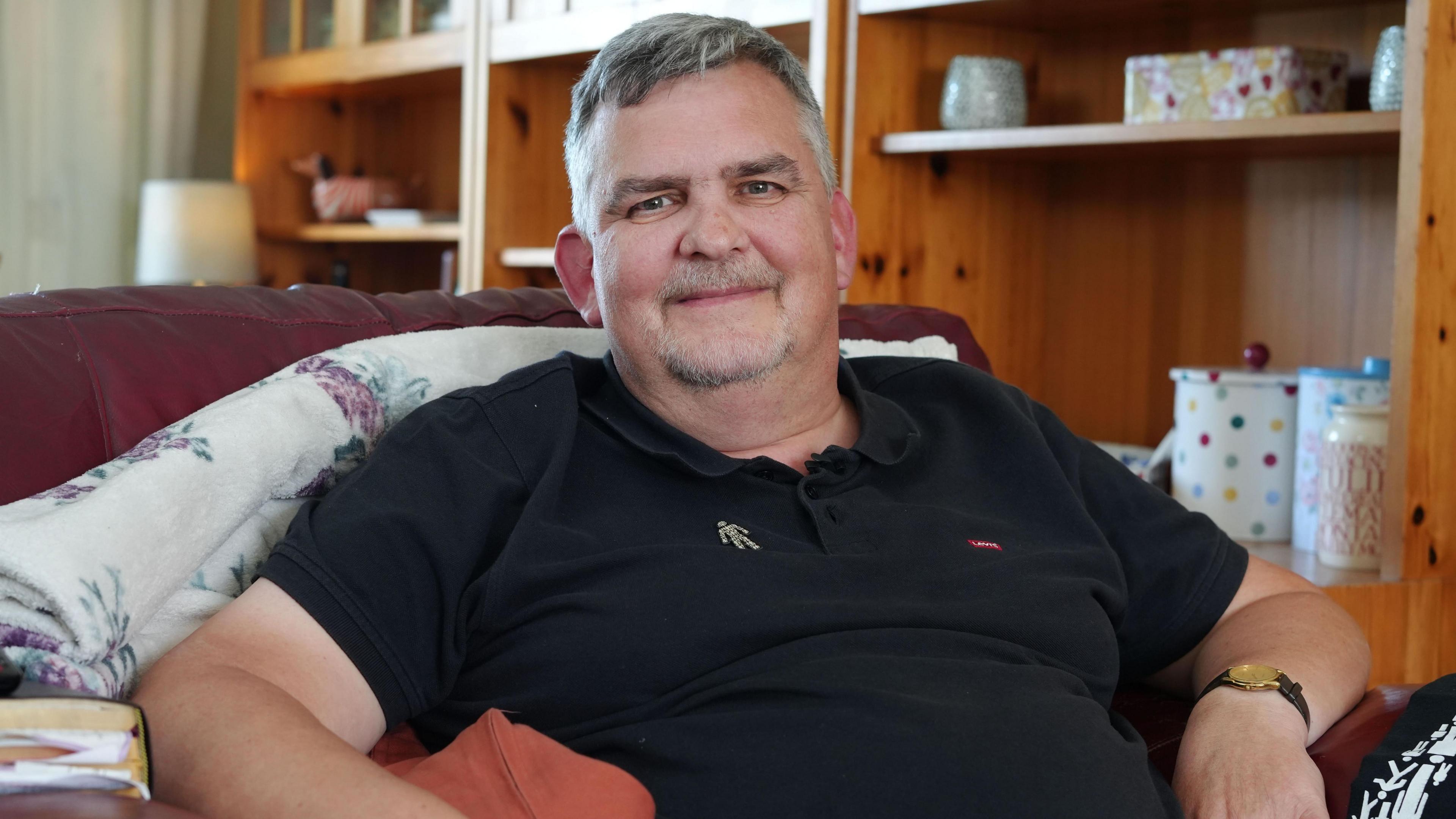 A man in a black polo shirt sits in a white armchair. He has grey hair and wears a Prostate Cancer UK badge.