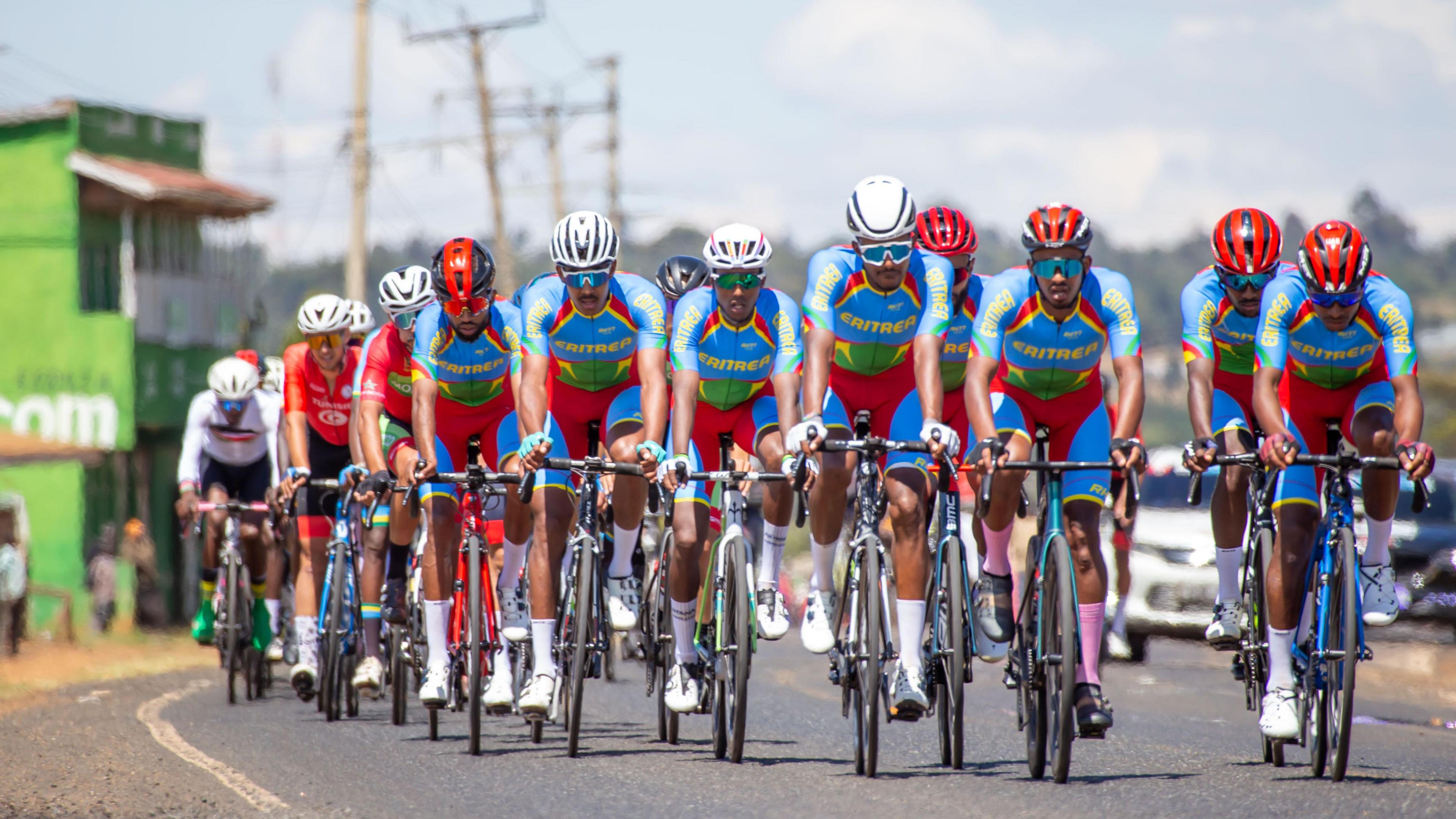 Eight Eritrean cyclists, wearing a blue, red, yellow and green kit, ride at the head of a peloton during a race on a road in Kenya