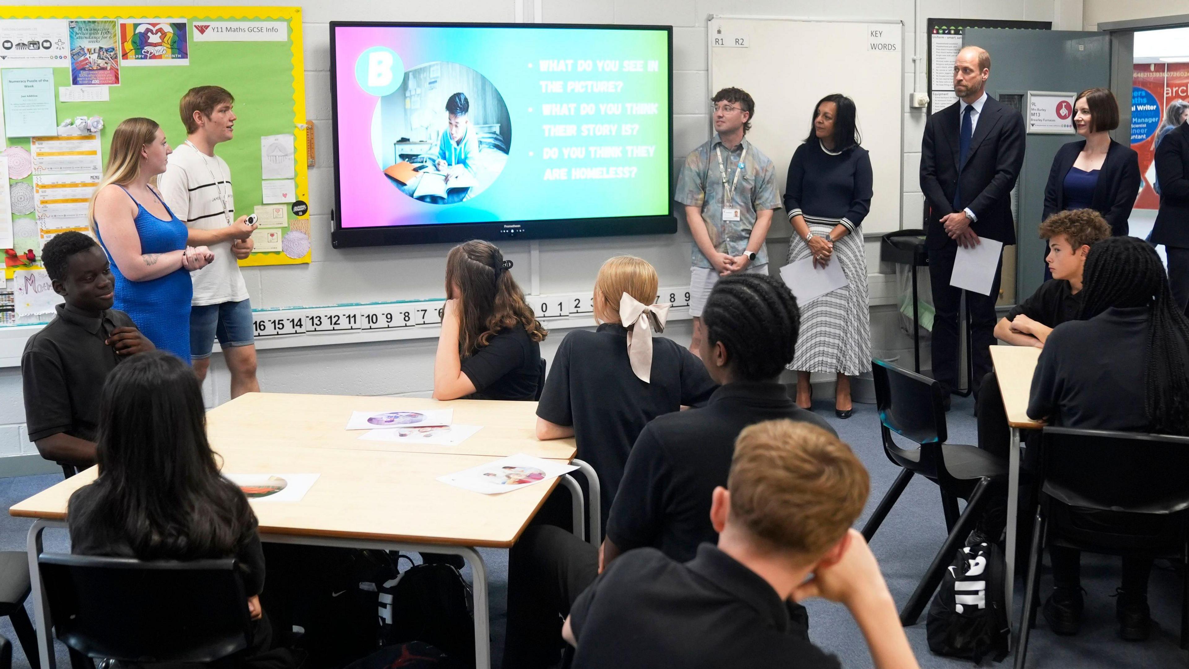 Prince William is stood in a classroom at Meadowhead School, surrounded by members of staff and students who are sat at their desks, watching a presentation on an electronic whiteboard.