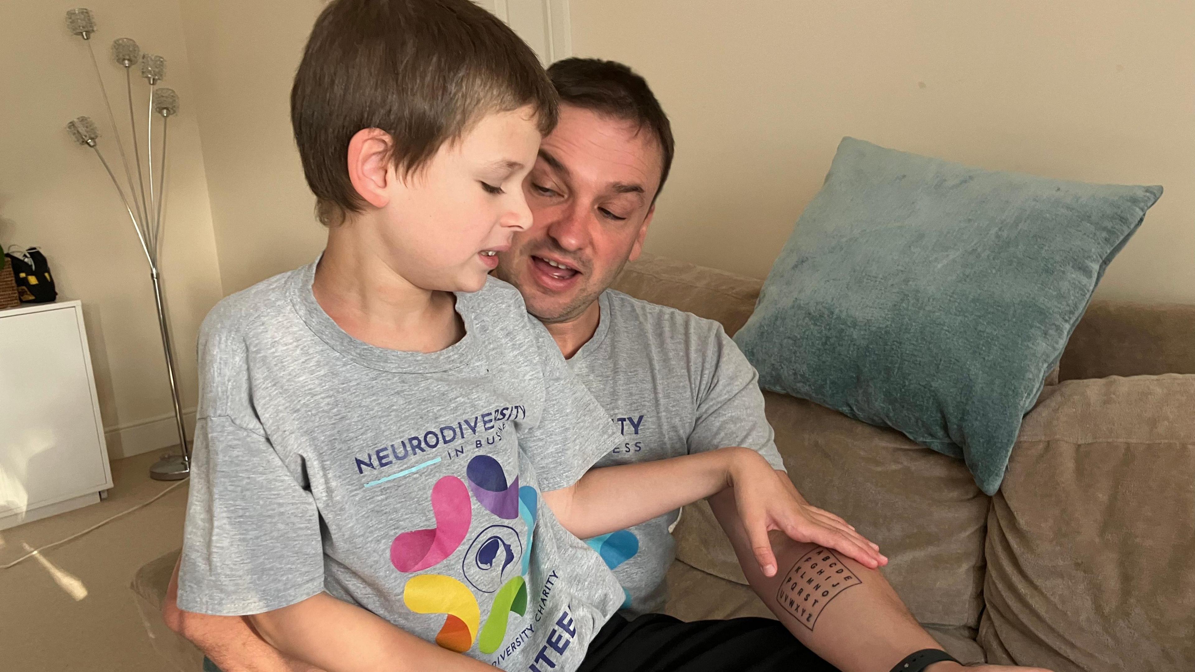 Joshie sitting on his dad's lap, both are wearing grey T-shirts and black trousers 