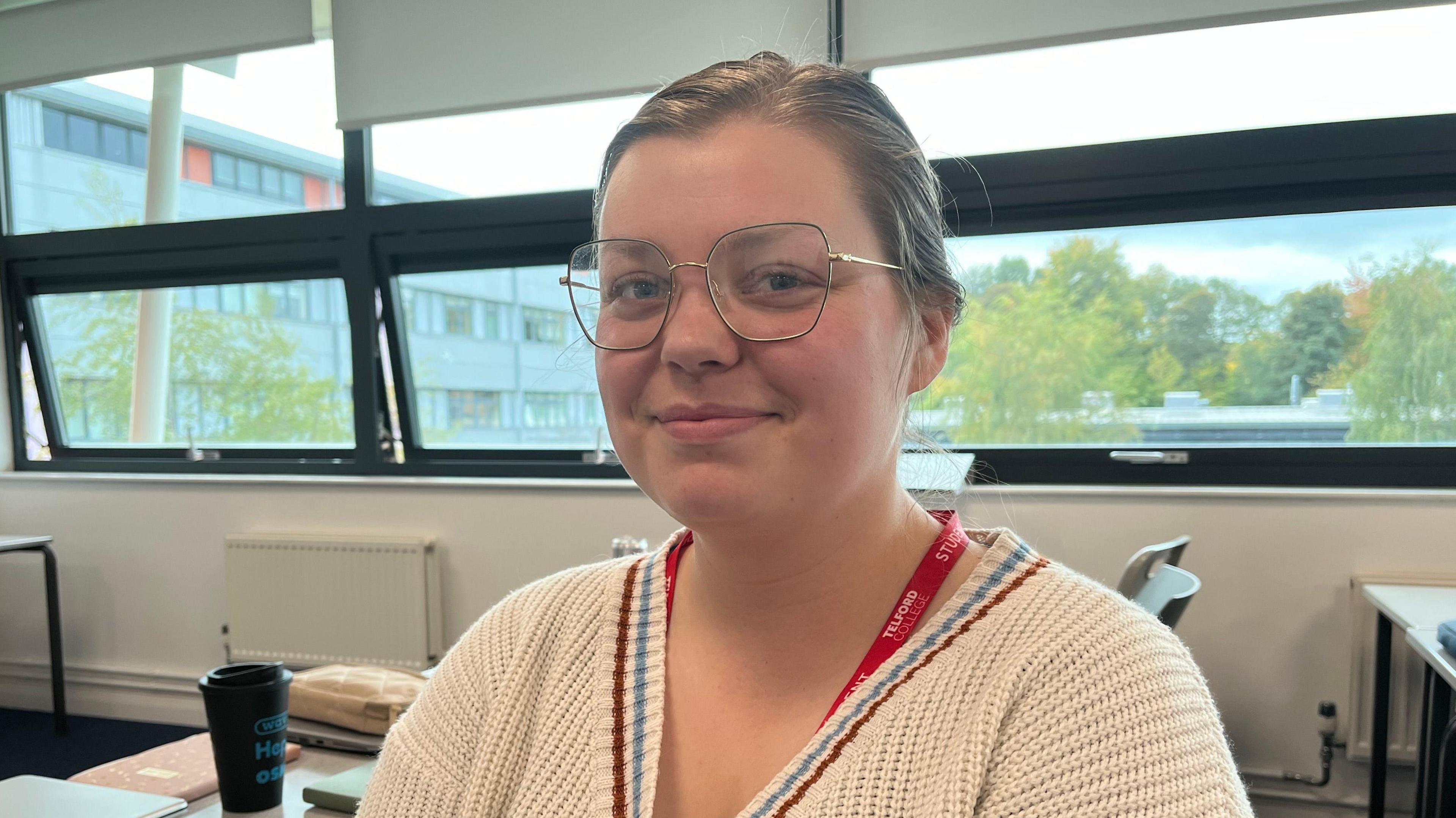 Megan pictured smiling slightly and looking into the camera. She's wearing a white cardigan and has a red Telford College lanyard on. Behind her is a large window looking out over the campus, which has lots of trees that are just starting to turn their leaves. Inside the classroom, there are white walls and grey desks. There's a black coffee cup on the desk directly behind Megan.