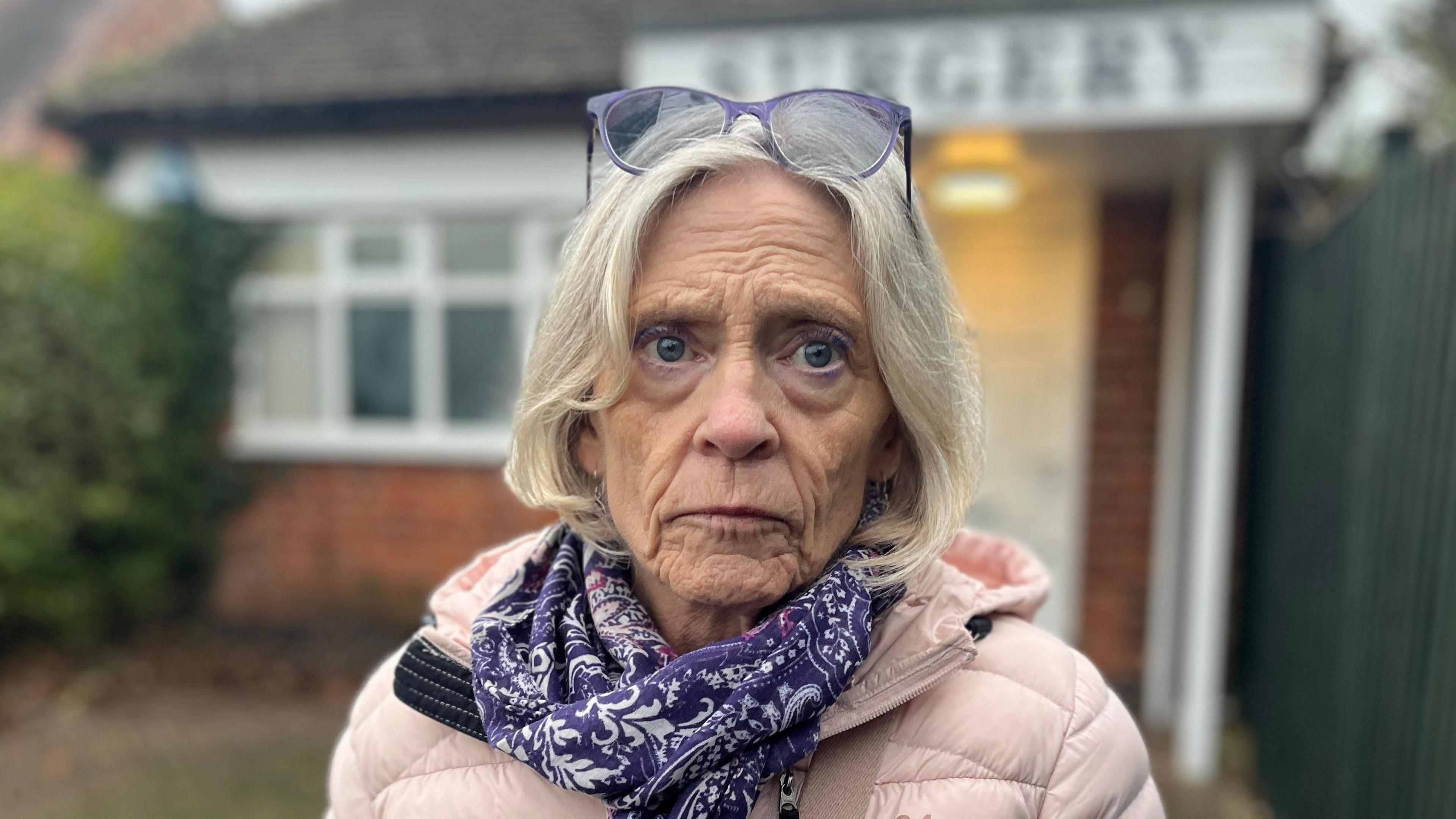 Anne Turner looking at the camera wearing a purple scarf and pink coat, with glasses on top of her head.