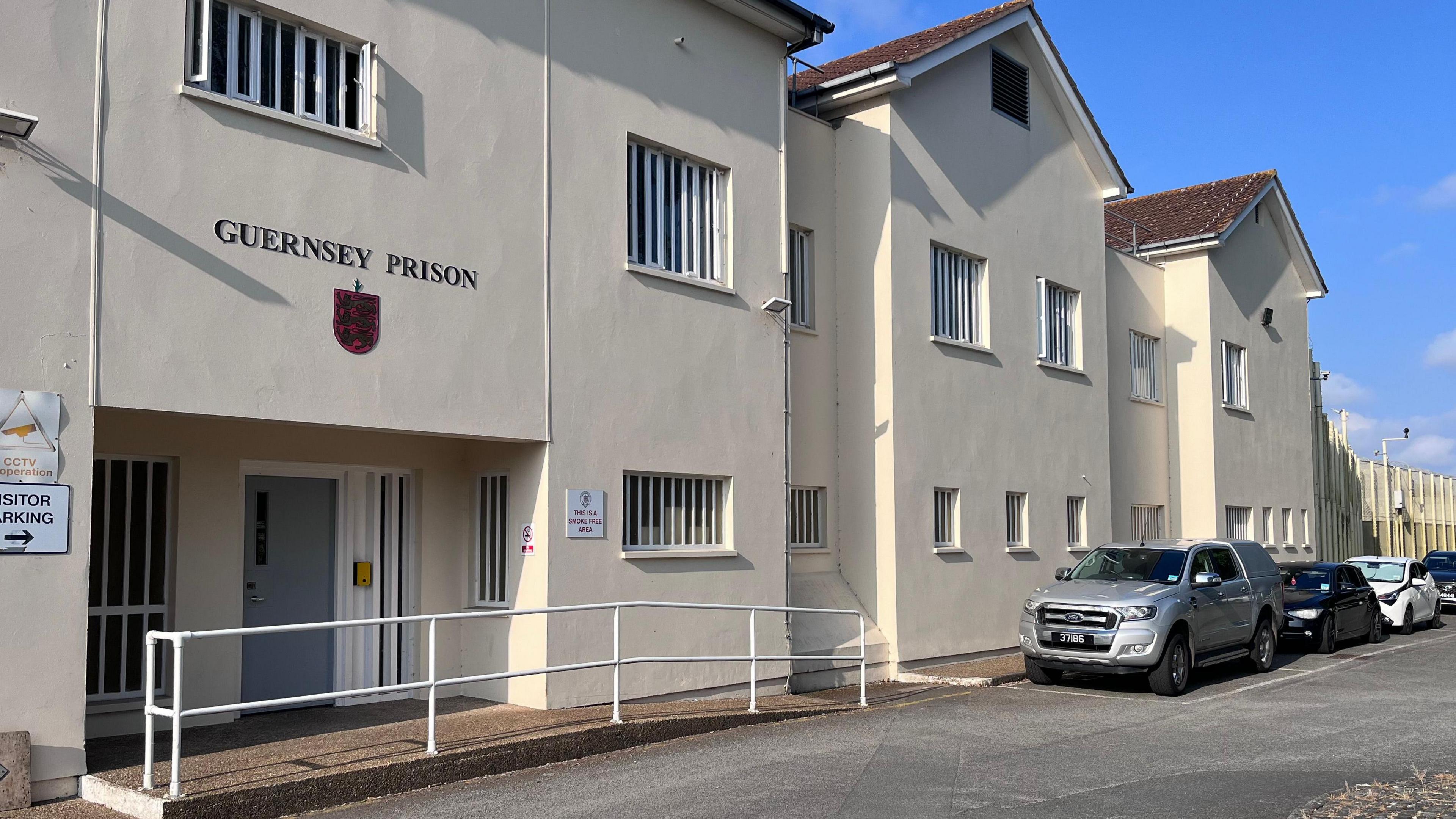 A large beige building with the words Guernsey Prison on the wall. In front of the building are a few cars parked.