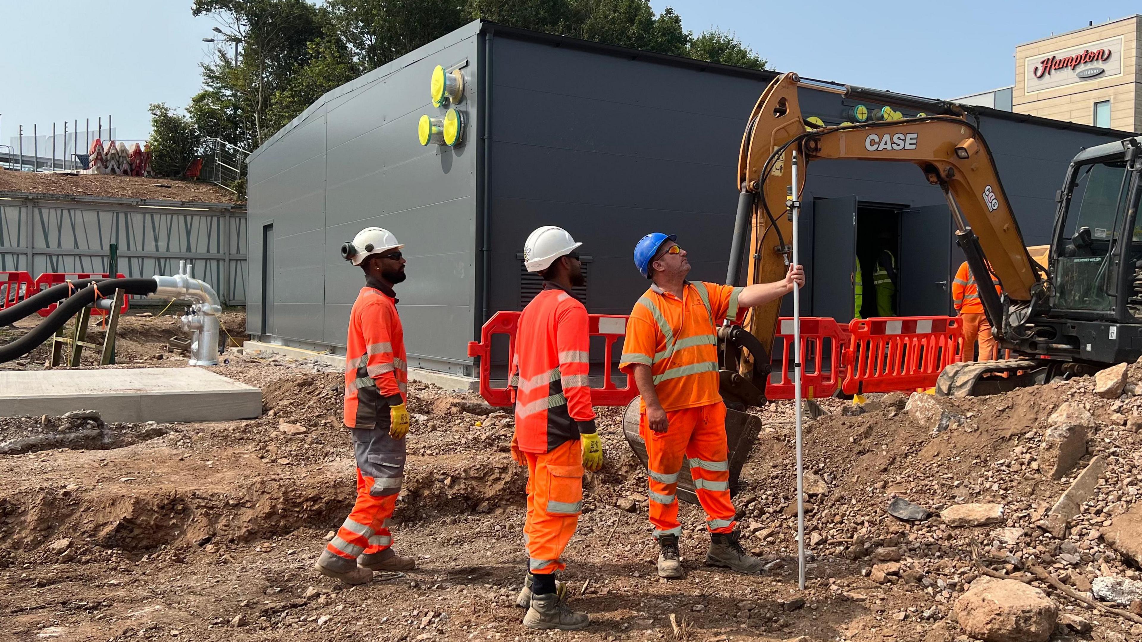 Workers in orange high vis outfits stood near yellow crane on rubble, with black shed in background.