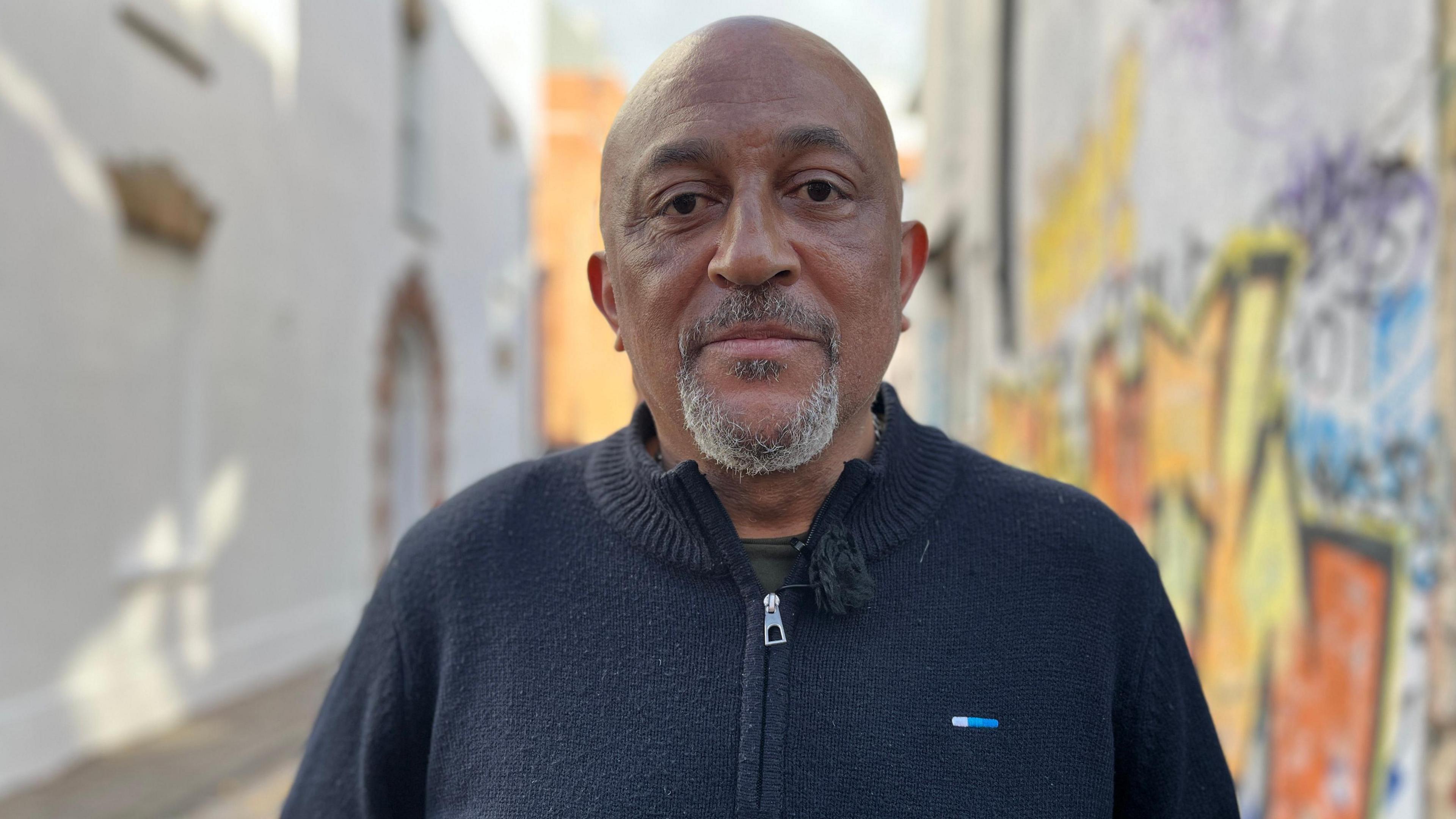 Desmond Brown looking at the camera. He is wearing a blue zip-up jumper. In the background blurred graffiti can be seen on a wall. 