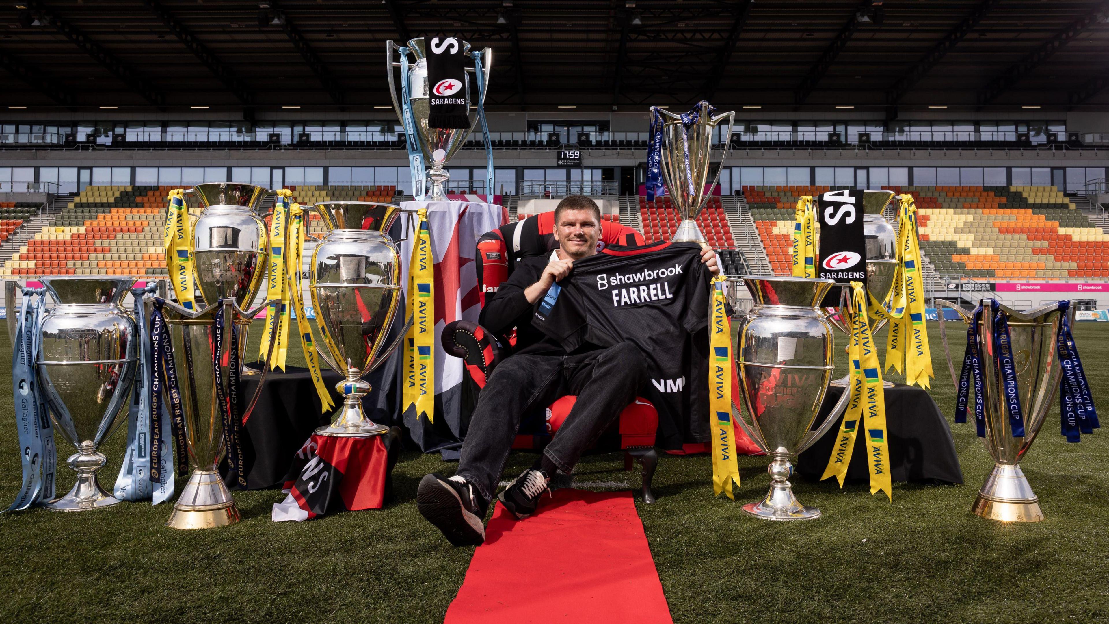 Owen Farrell sits on a chair in the middle of the StoneX Stadium pitch holding a shirt that says 'Farrell' with a red carpet in front of him and nine trophies placed around him 