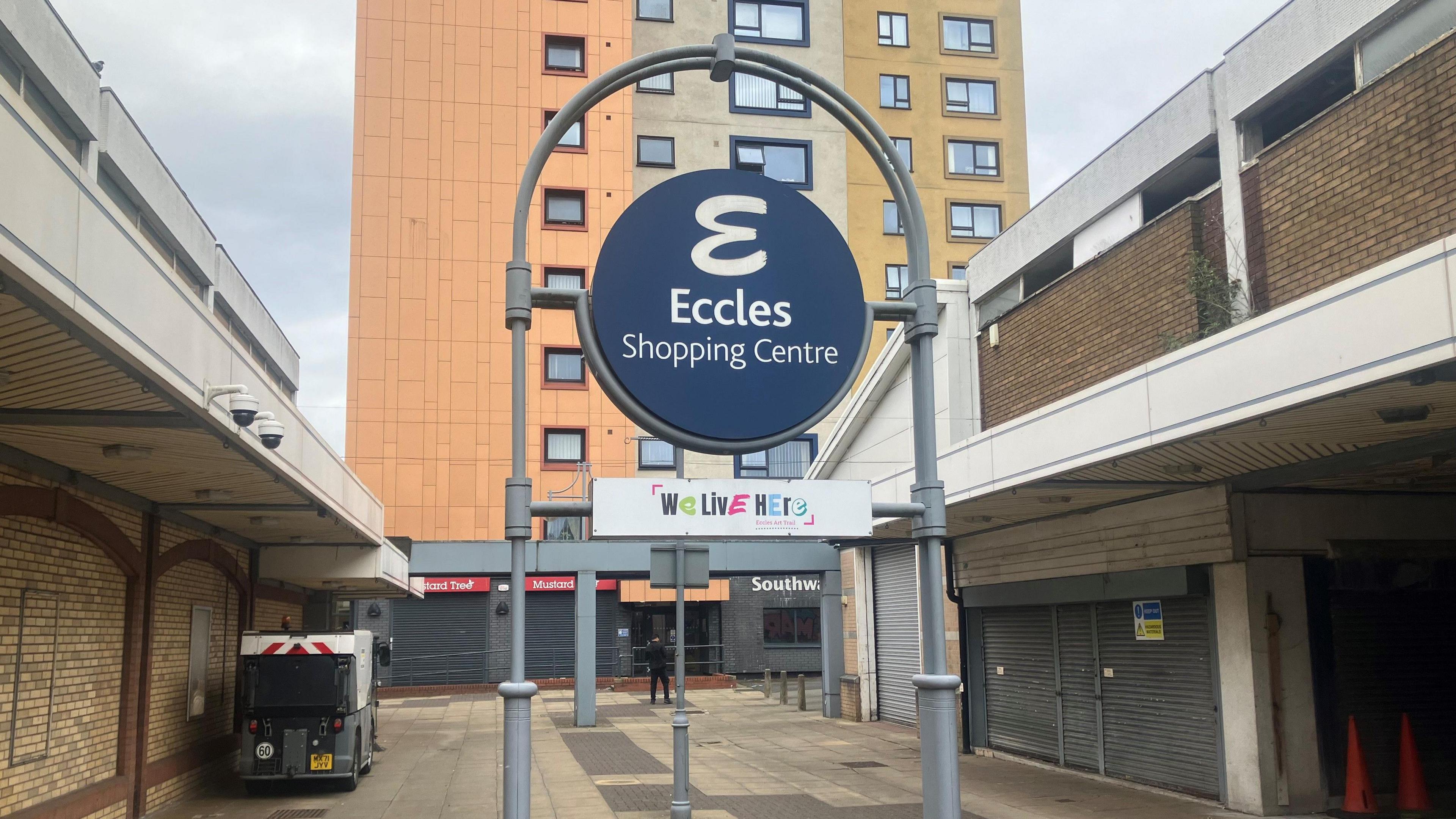 The entrance to Eccles Shopping Centre with a large sign in the precinct. In the background and to the right hand side are shuttered up shops. There is one lone person in the area, otherwise it is empty.