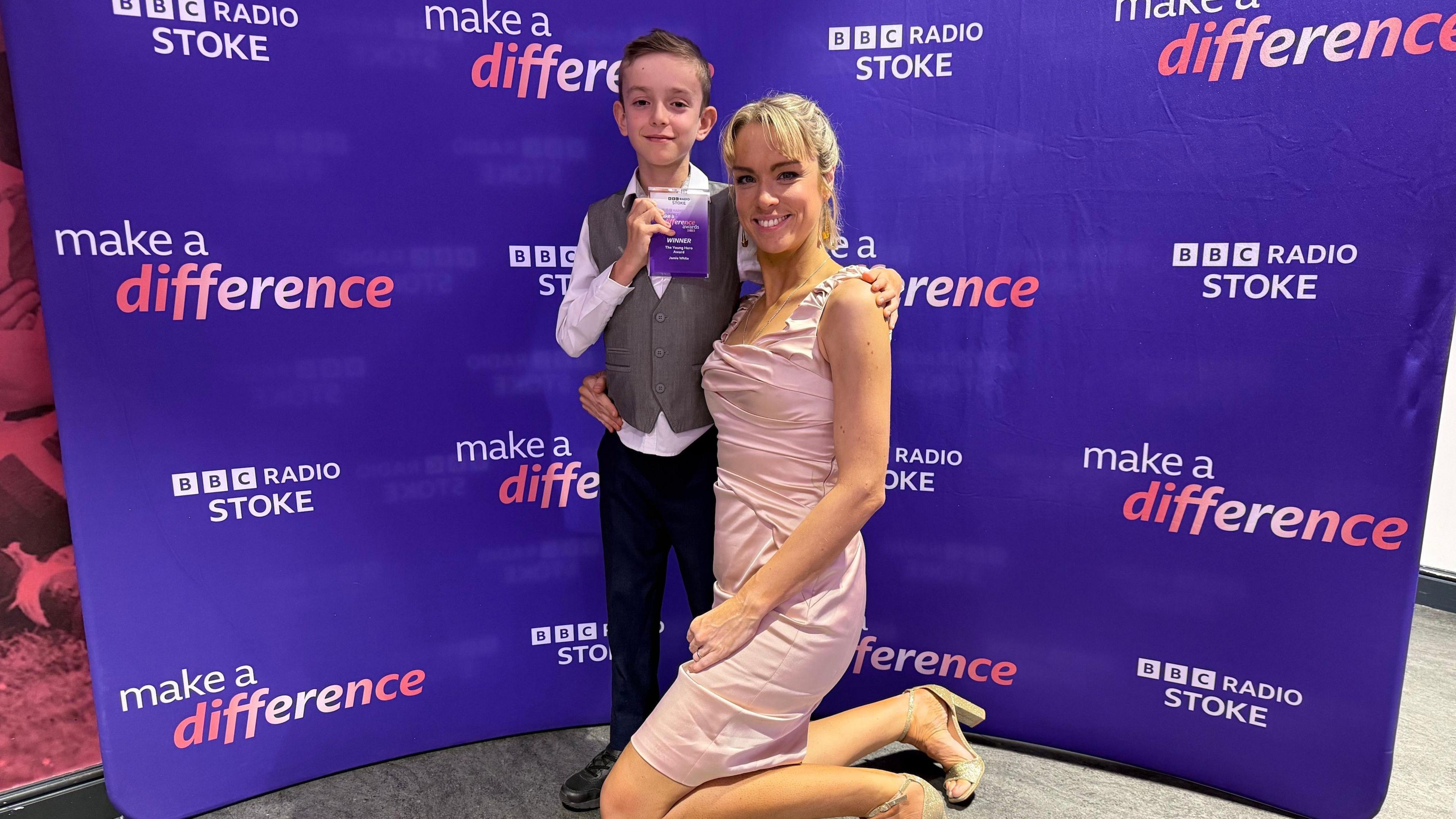 Jamie and his mum. Jamie is wearing a grey waistcoat and white shirt with black trousers. His mum is wearing a pink dress. Jamie is standing while his mum is kneeling in front of a purple BBC Radio Stoke Make A Difference step and repeat board.