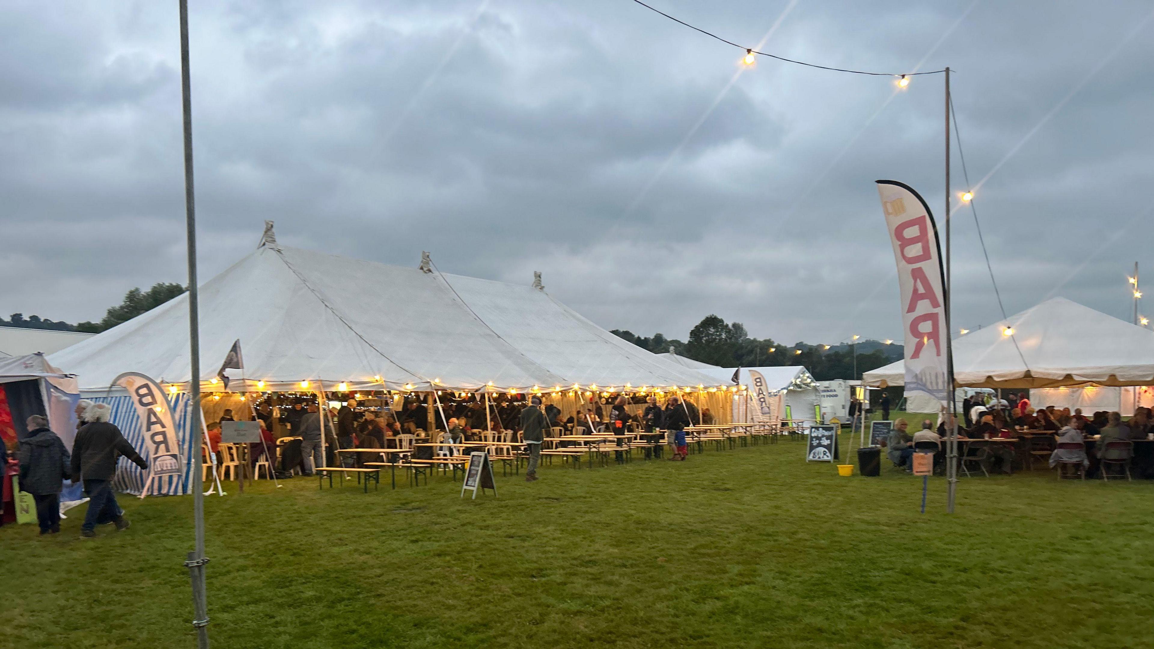 A white gazebo in the middle of a field with fairy lights draped across the edge. There a large flags that read "beer" in red lettering 