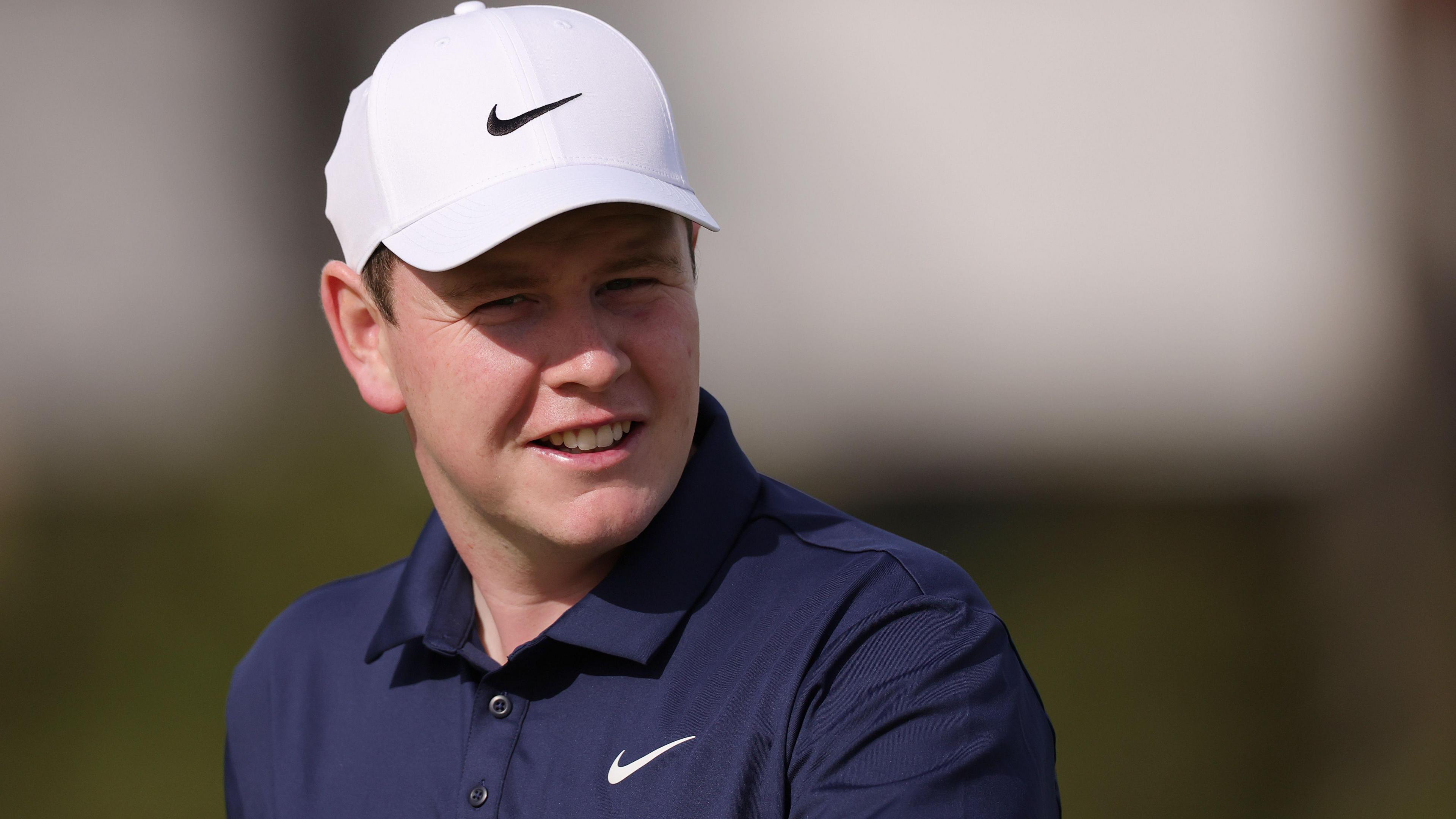 Head and shoulders shot of a smiling Robert MacIntyre. He is wearing a dark blue polo shirt and white baseball cap.
