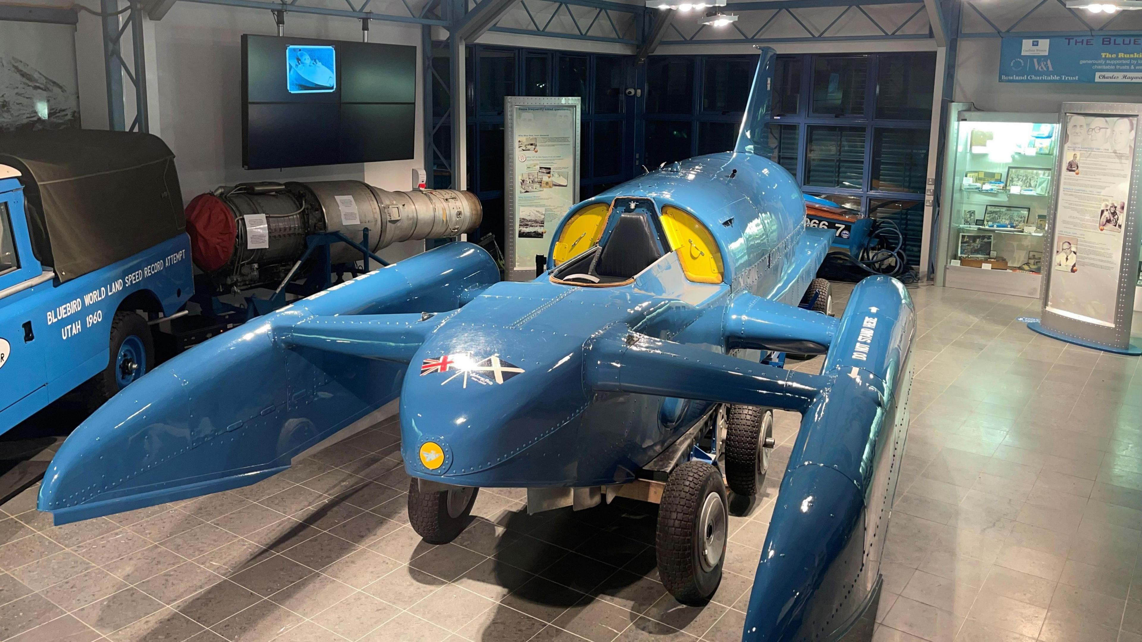 Bluebird K7 on display at the Ruskin Museum in Coniston. The blue craft with yellow details has a single seat for the pilot at the front. It has flags of the UK and Scotland painted at the front.
