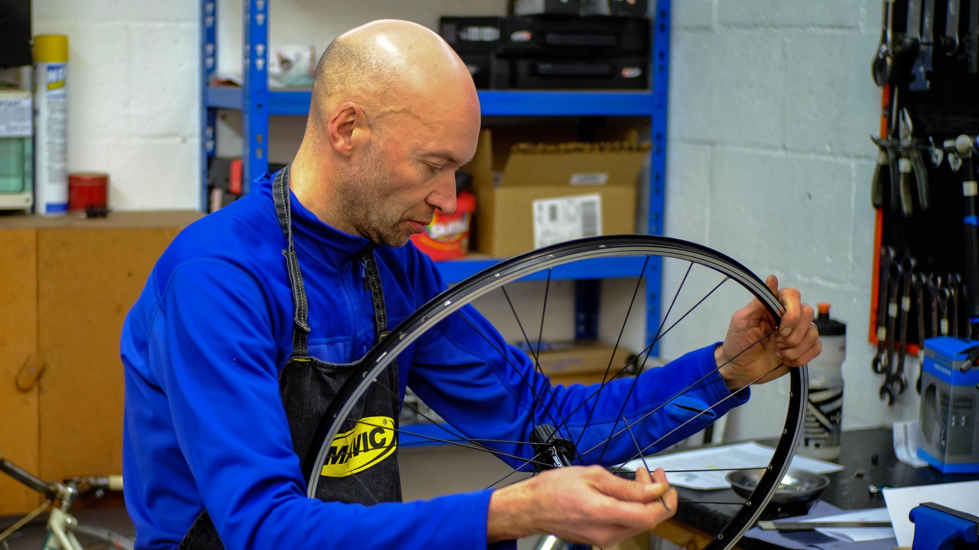 A man wearing a blue top and black apron holding a bicycle wheel in a workshop 